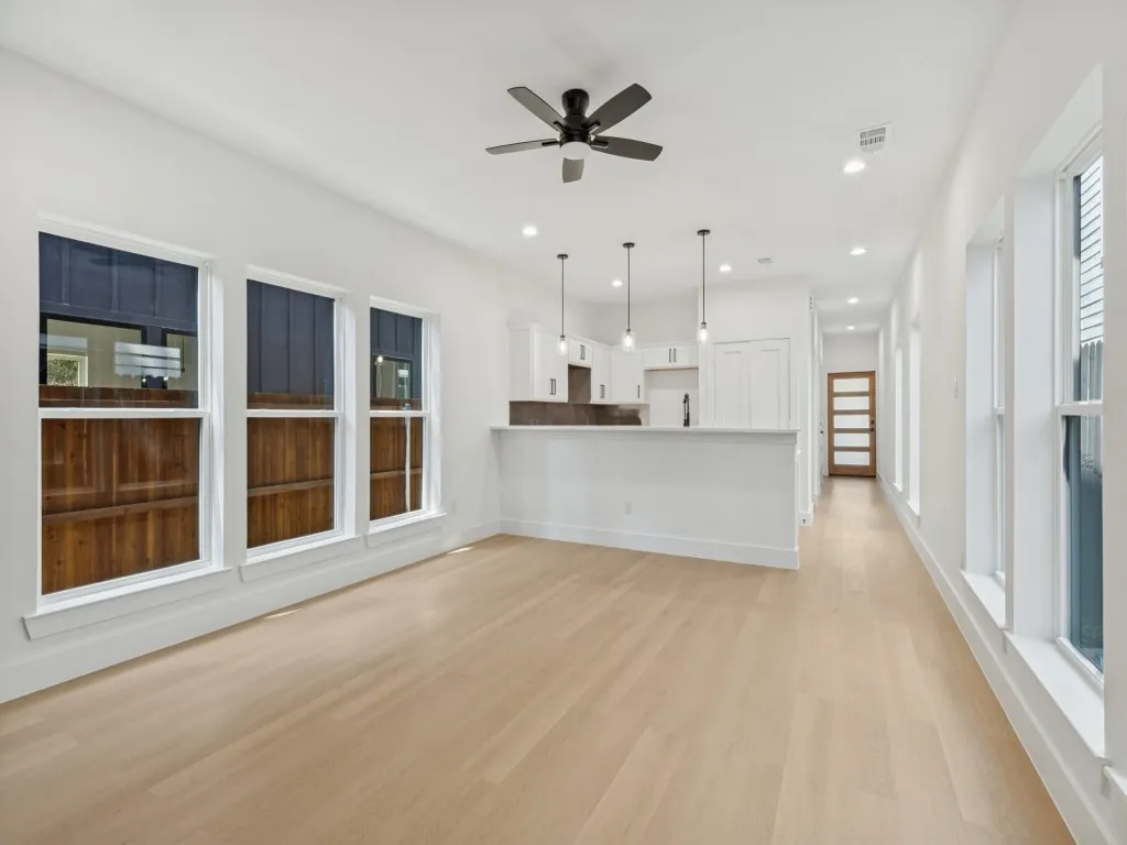 Open-concept living area featuring light wood-finish flooring, multiple windows, a modern ceiling fan, recessed lighting, and a kitchen with white cabinetry
