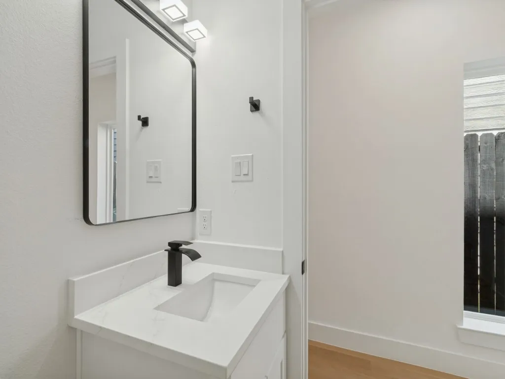 White vanity with a rectangular sink, matte black faucet, and a framed mirror with integrated lighting