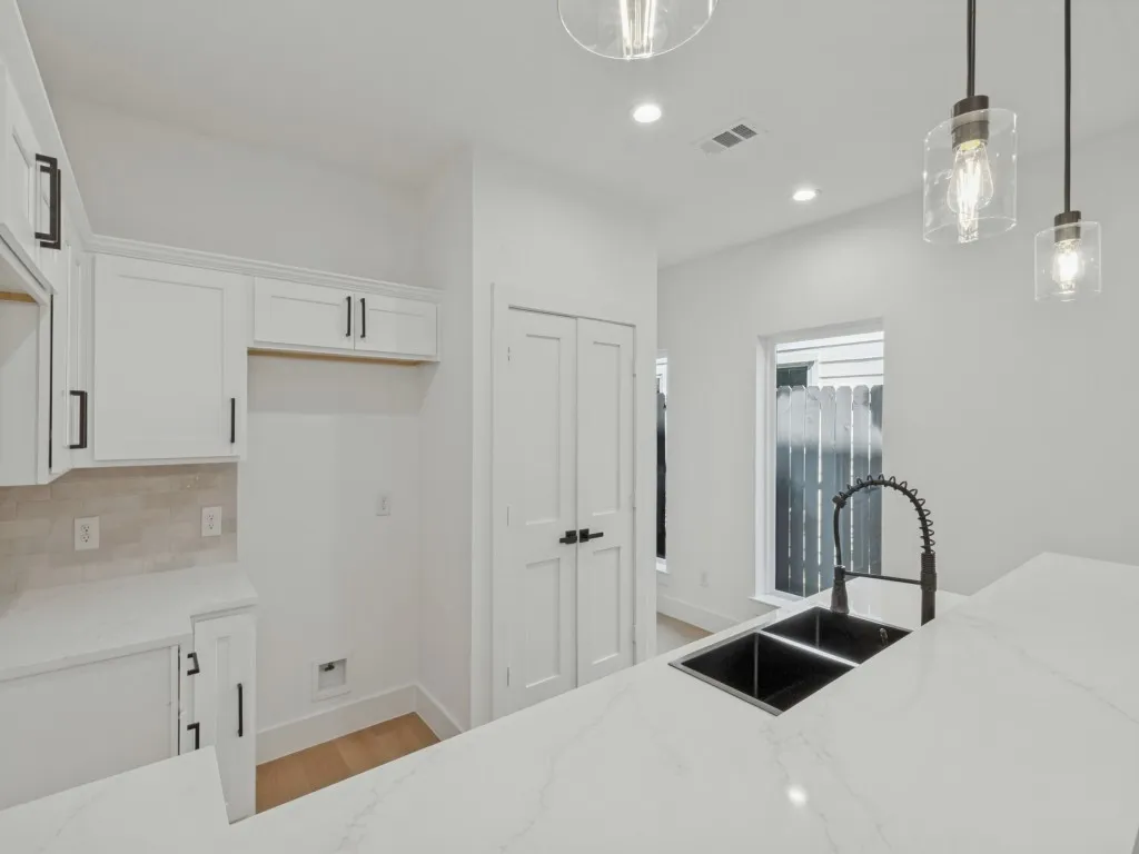 White shaker cabinetry with matte black hardware, stone-finish countertops, dual-basin sink with gooseneck faucet, light wood-finish flooring, and pendant lighting