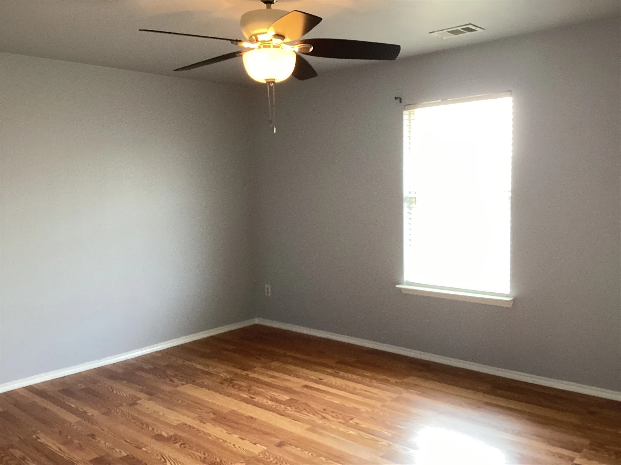 Wood-finish flooring, light gray walls, single window with blinds, and a ceiling fan with integrated lighting