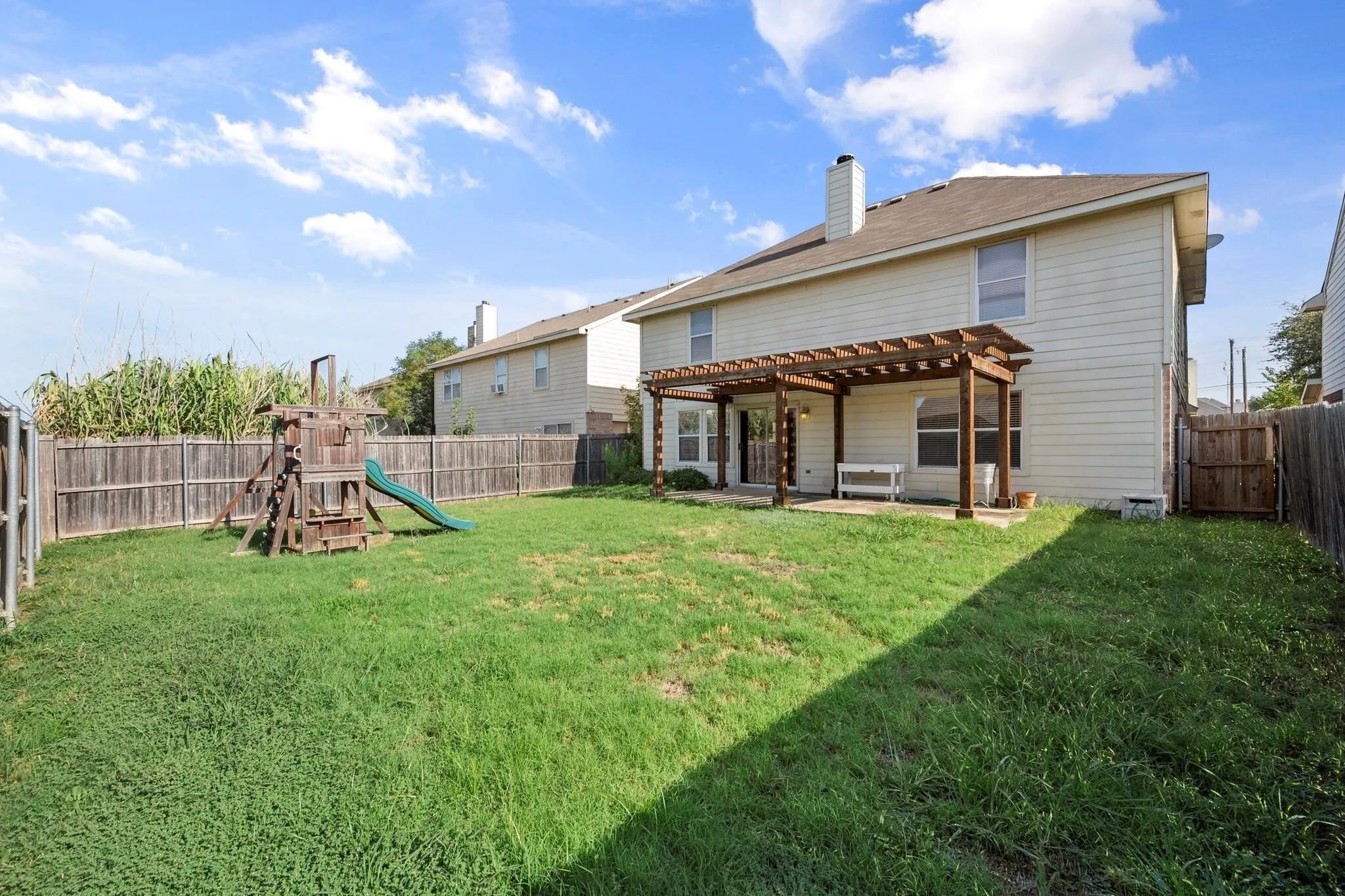 Expansive backyard featuring a wood privacy fence, a two-story residence with light-colored siding, and a wooden pergola covering a patio area