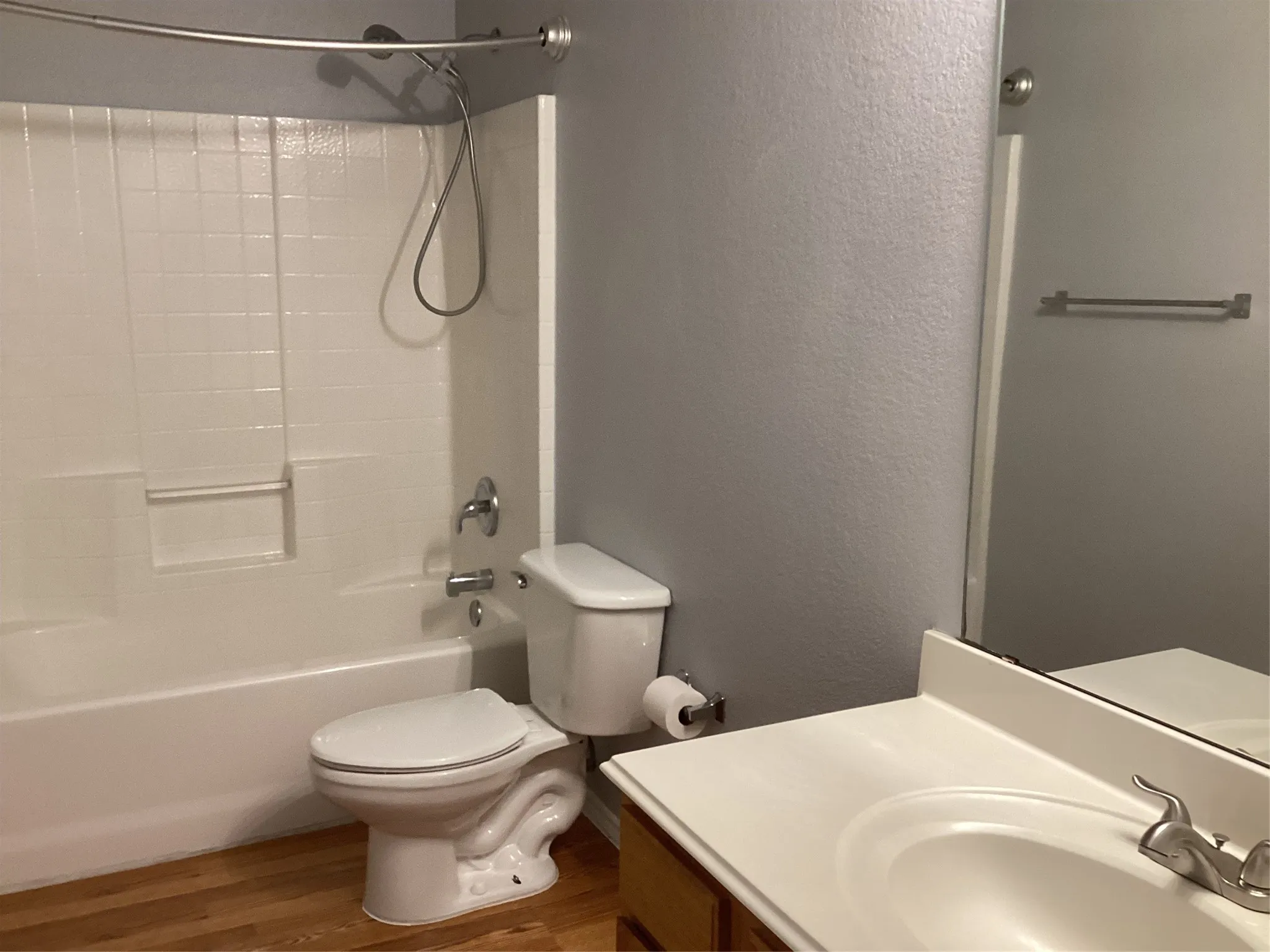 White bathtub with integrated shower, white toilet, wood-finish flooring, and a vanity with an integrated sink