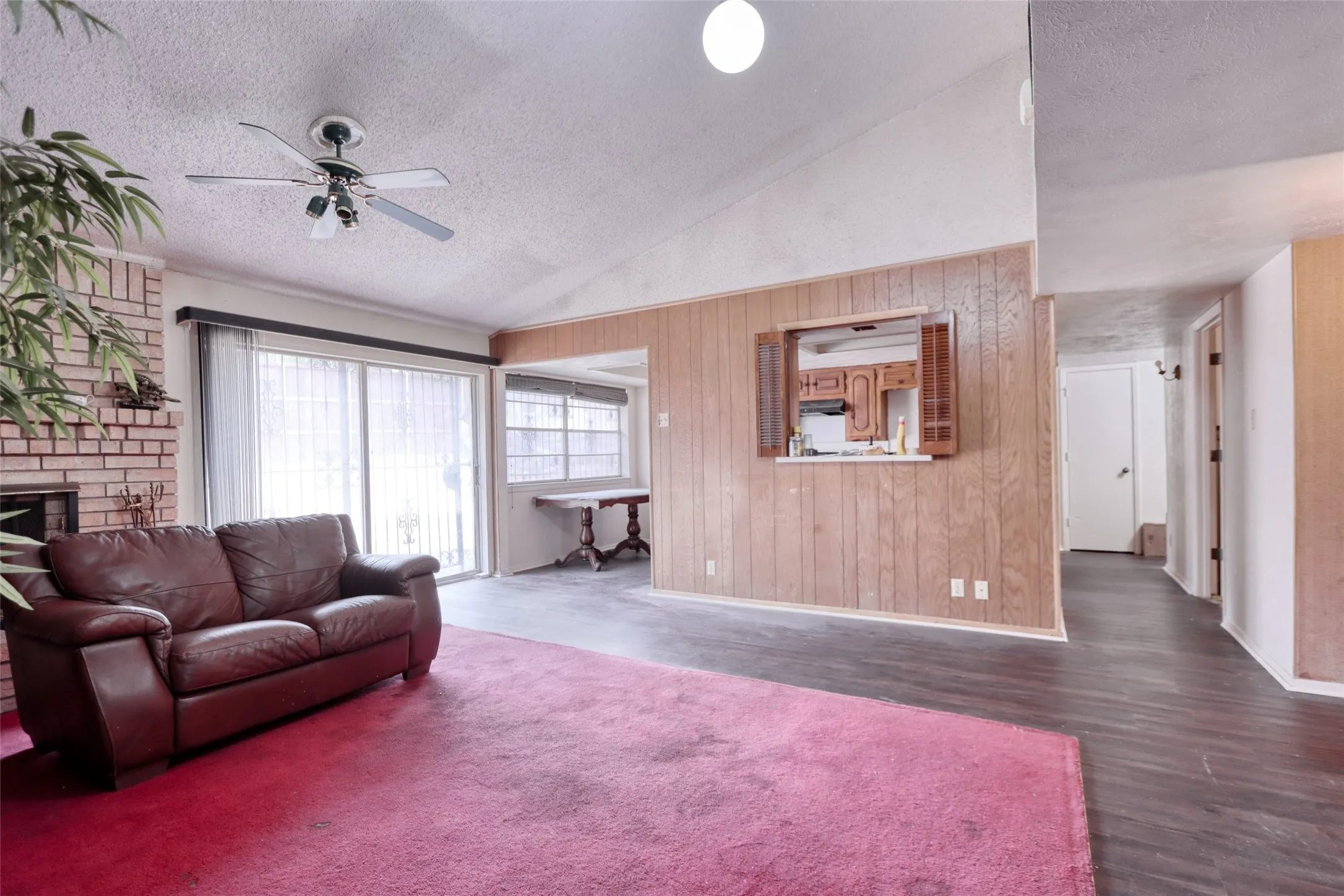 Brick fireplace with hearth, ceiling fan, wood-panel accent wall, pass-through window with cabinetry, and sliding glass door