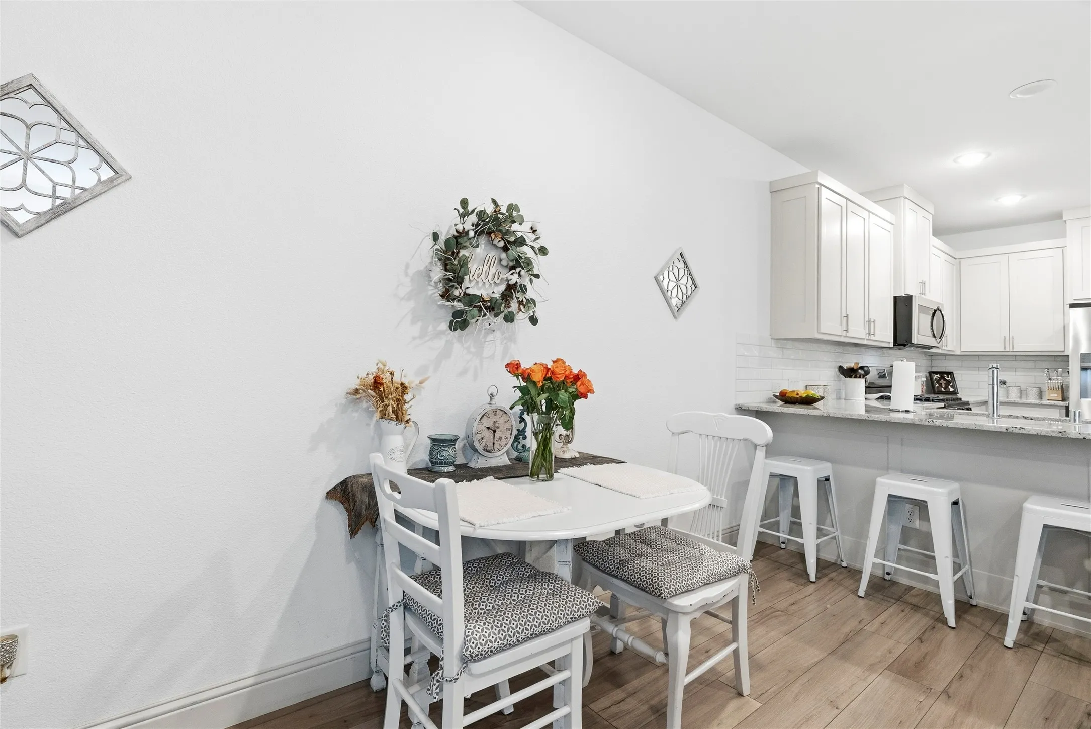Open-concept dining and kitchen area featuring a white ladder-back dining table, wood-finish flooring throughout, white cabinetry with stainless steel appliances, and a white quartz countertop island with seating for three