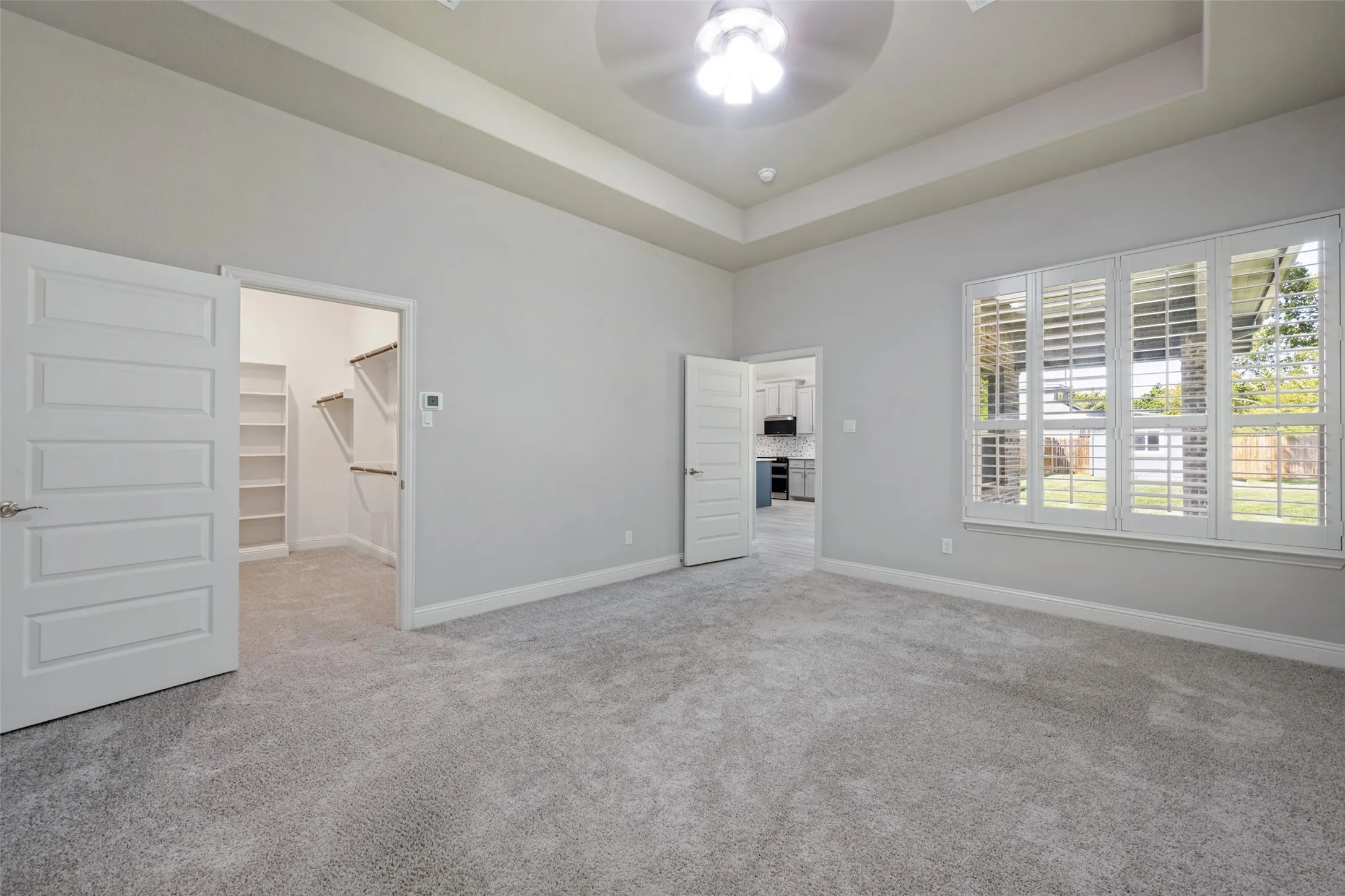 Recessed tray ceiling with ceiling fan, plush gray carpet, a walk-in closet with built-in shelving, and a large window with plantation shutters are features of the primary bedroom.
