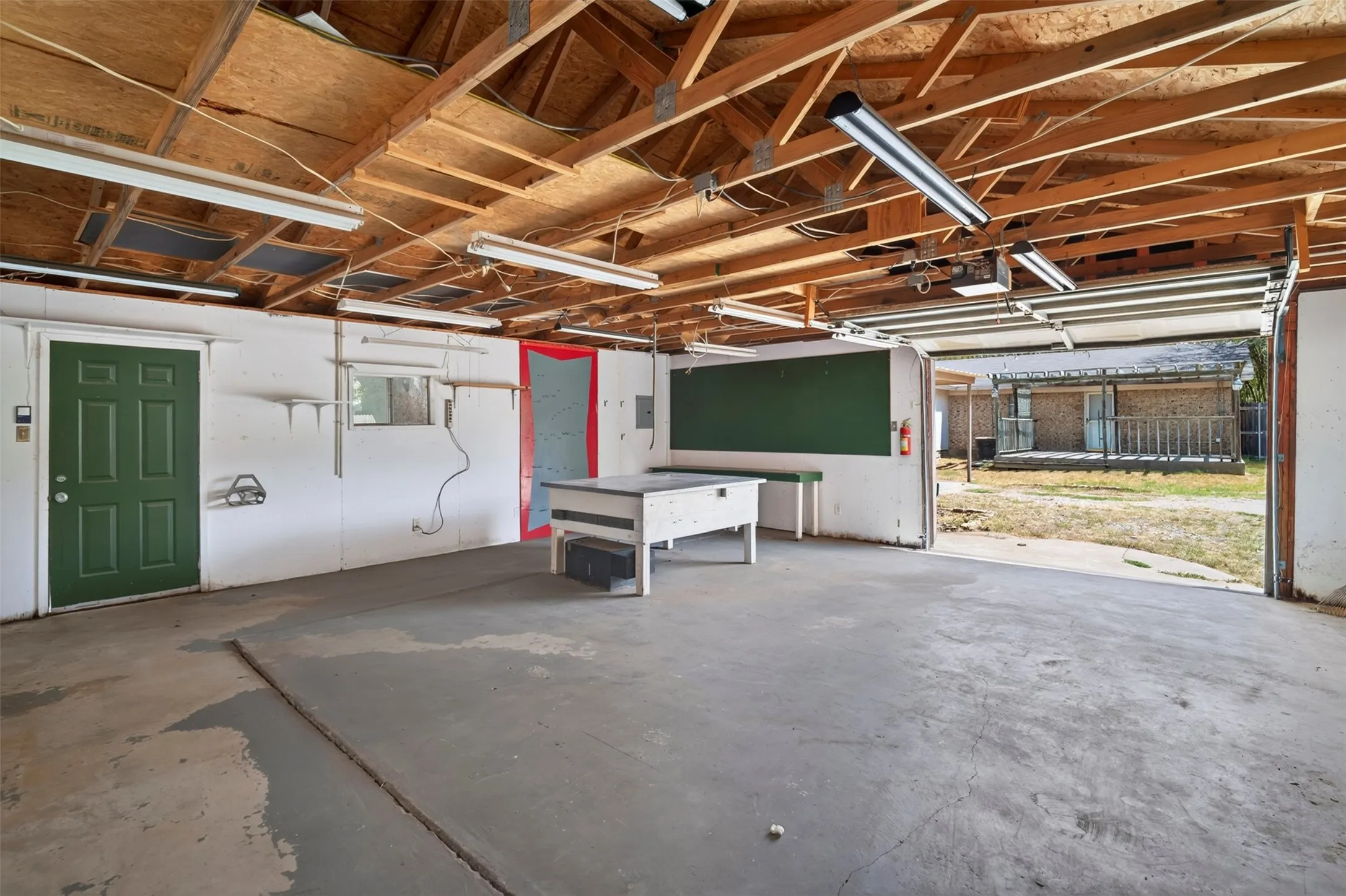 Open garage space with exposed wood beam ceiling, concrete flooring, fluorescent lighting, and a green painted door