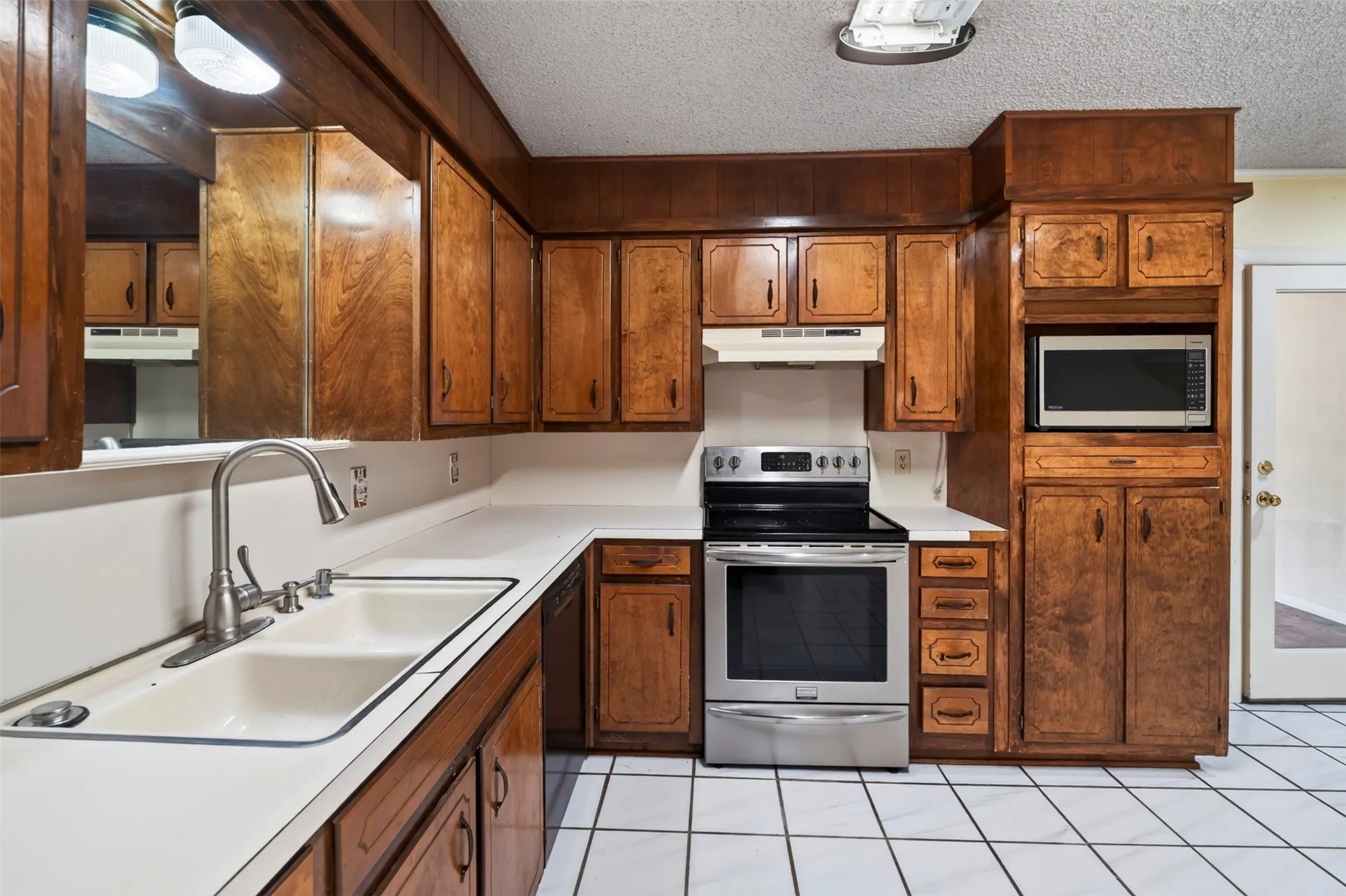 Wood-finish cabinetry, stainless steel range, built-in microwave, dual basin sink, and white tiled flooring