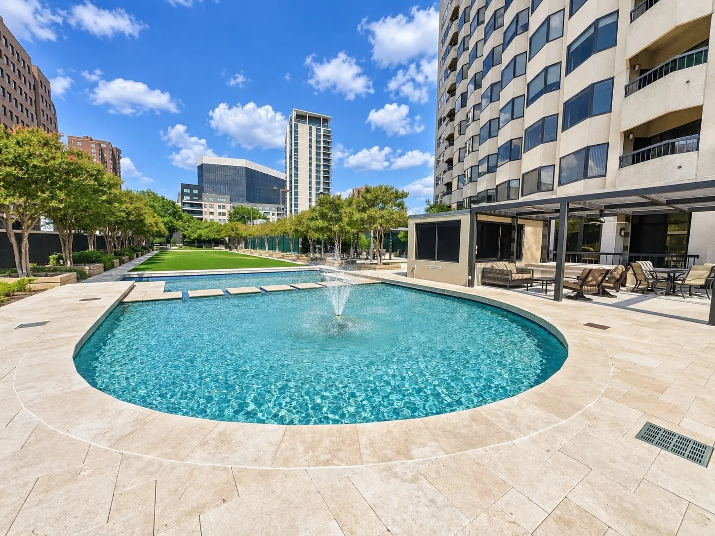 Resort-style pool with a fountain feature, expansive paved patio, multiple outdoor seating areas, lush green lawn, and mature landscaping