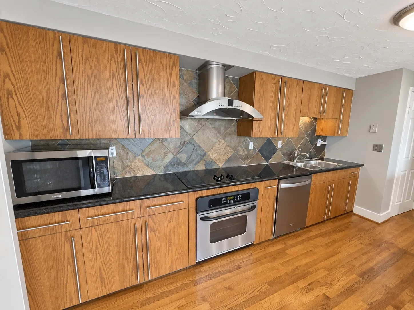 Kitchen featuring wood-finish cabinetry, stainless steel appliances, black countertops, a tile backsplash, and wood-finish flooring