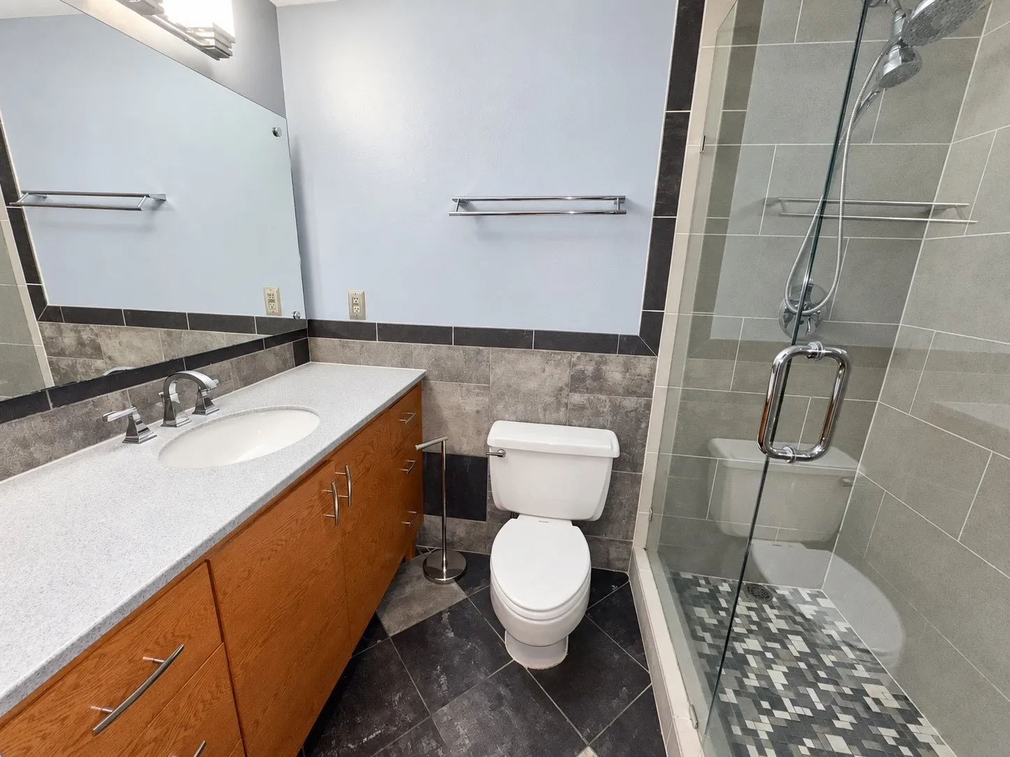 Glass-enclosed shower with a mosaic tile floor and integrated shower head, vanity with a light-toned countertop and undermount sink, wood-finish cabinetry, and tiled walls