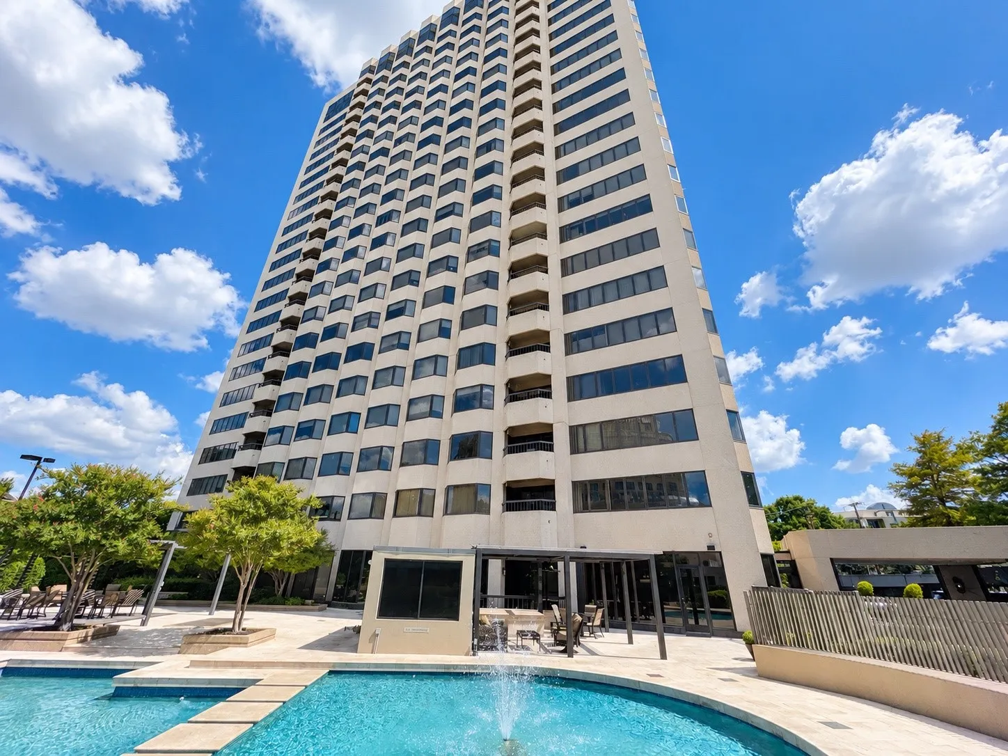 High-rise building with a tiered facade, featuring a multi-level pool with a fountain and integrated stepping stones, a covered outdoor lounge area, and mature landscaping