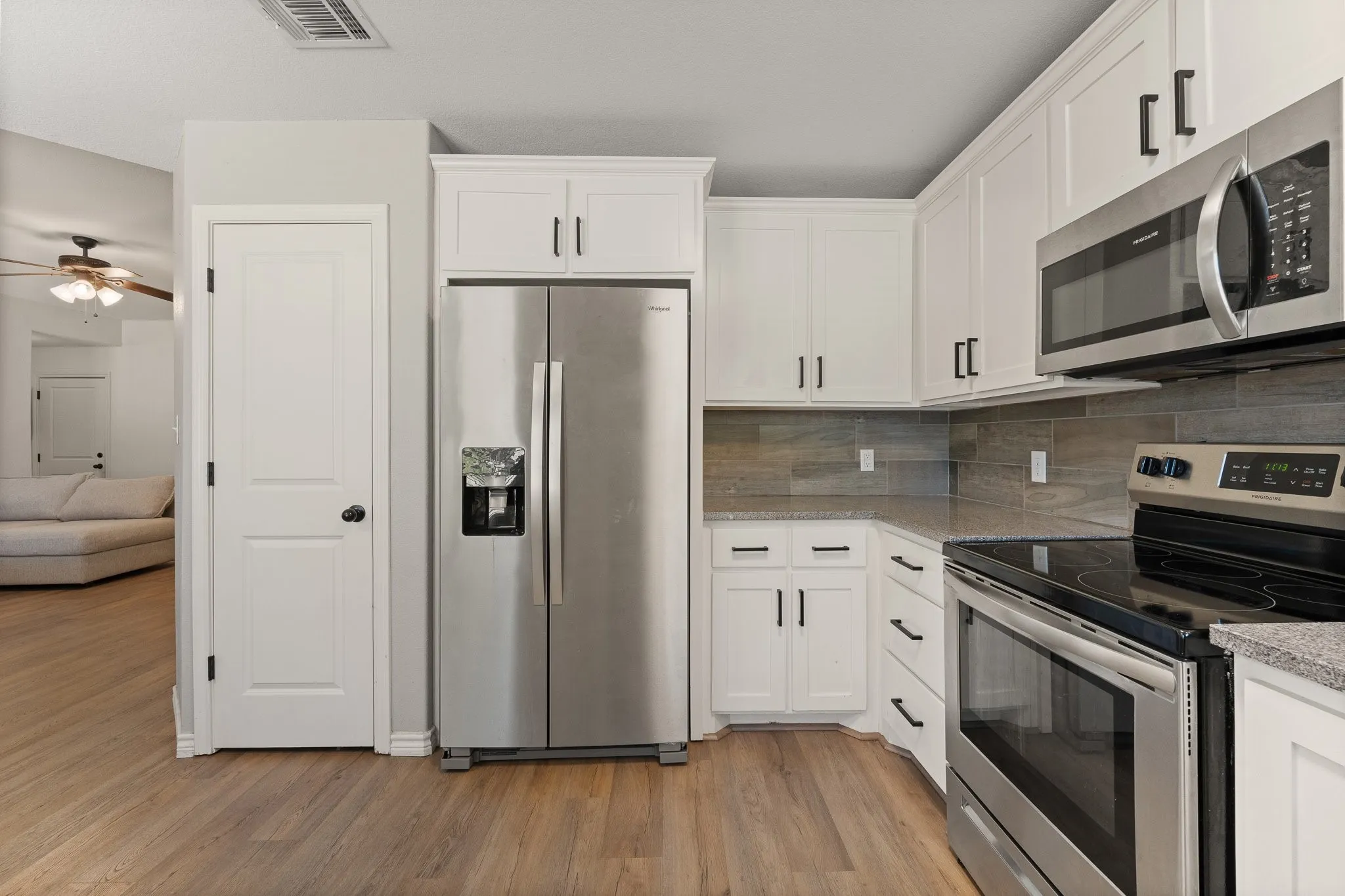 White shaker cabinetry, stainless steel appliances, wood-finish flooring, light-toned countertops, and a tiled backsplash