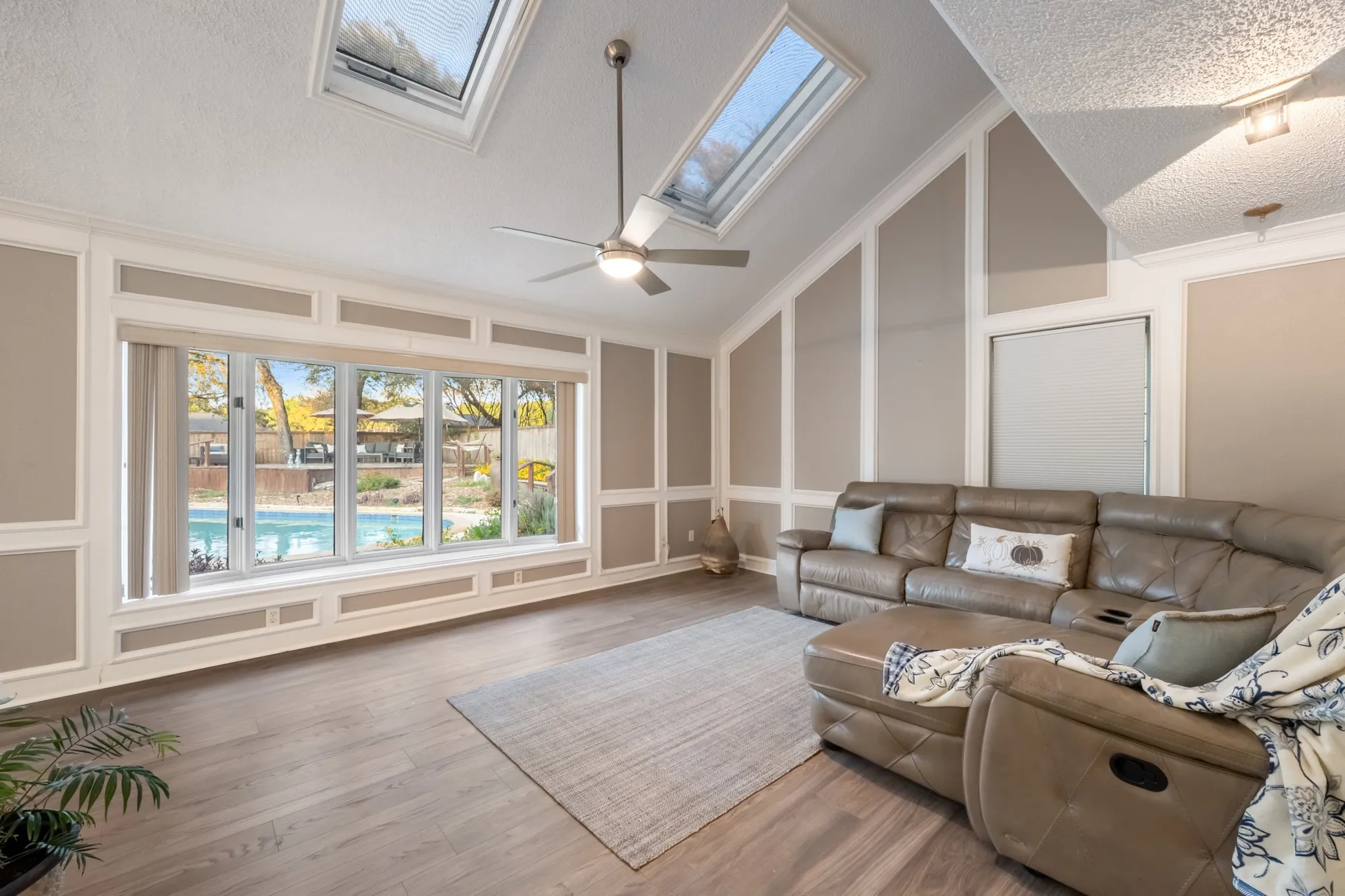 Vaulted ceiling with dual skylights, wood-finish flooring, accent wall paneling, expansive window overlooking a pool, and a ceiling fan