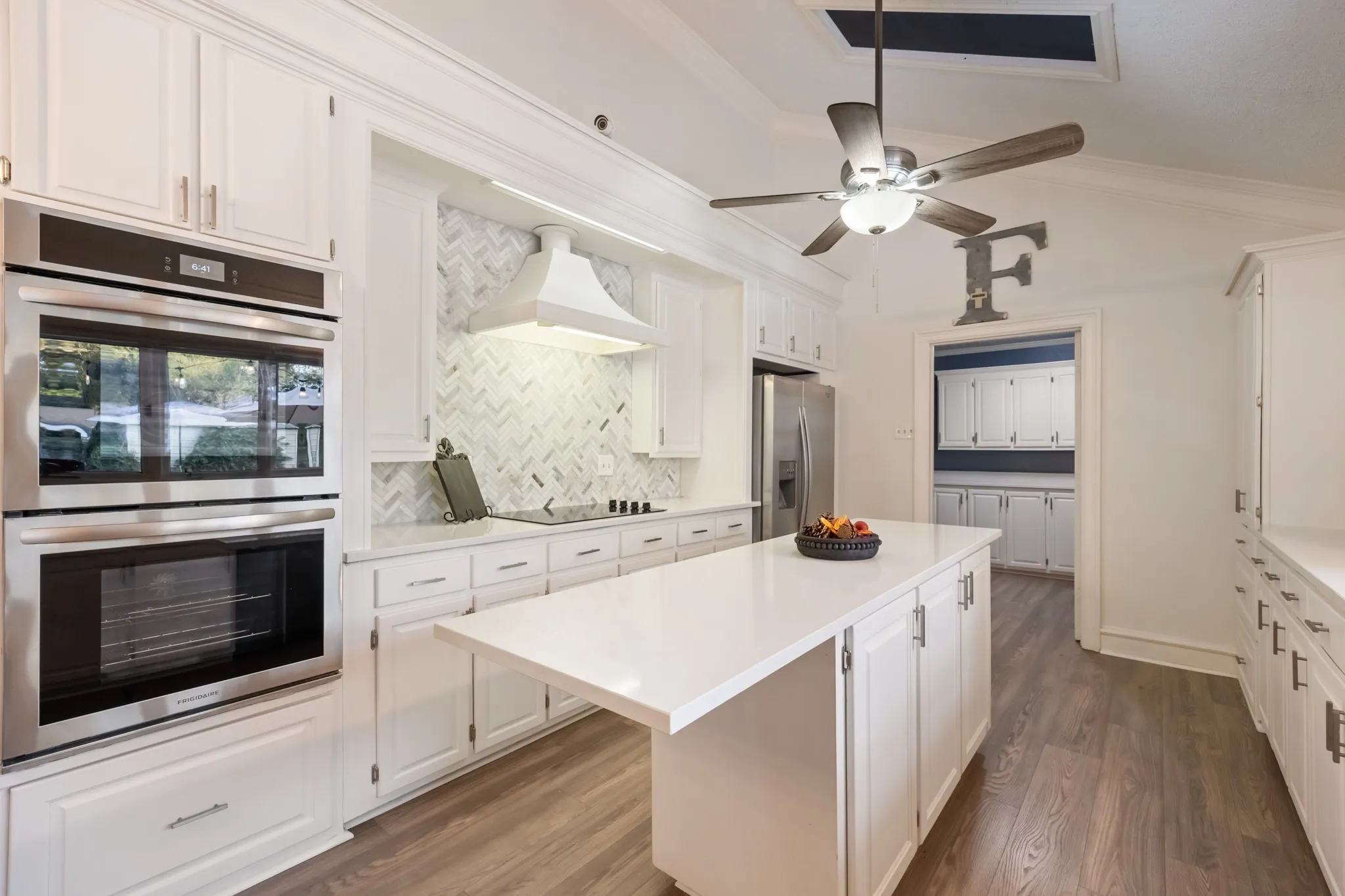White cabinetry, light wood-finish flooring, white countertops, built-in double oven, and a ceiling fan with integrated lighting