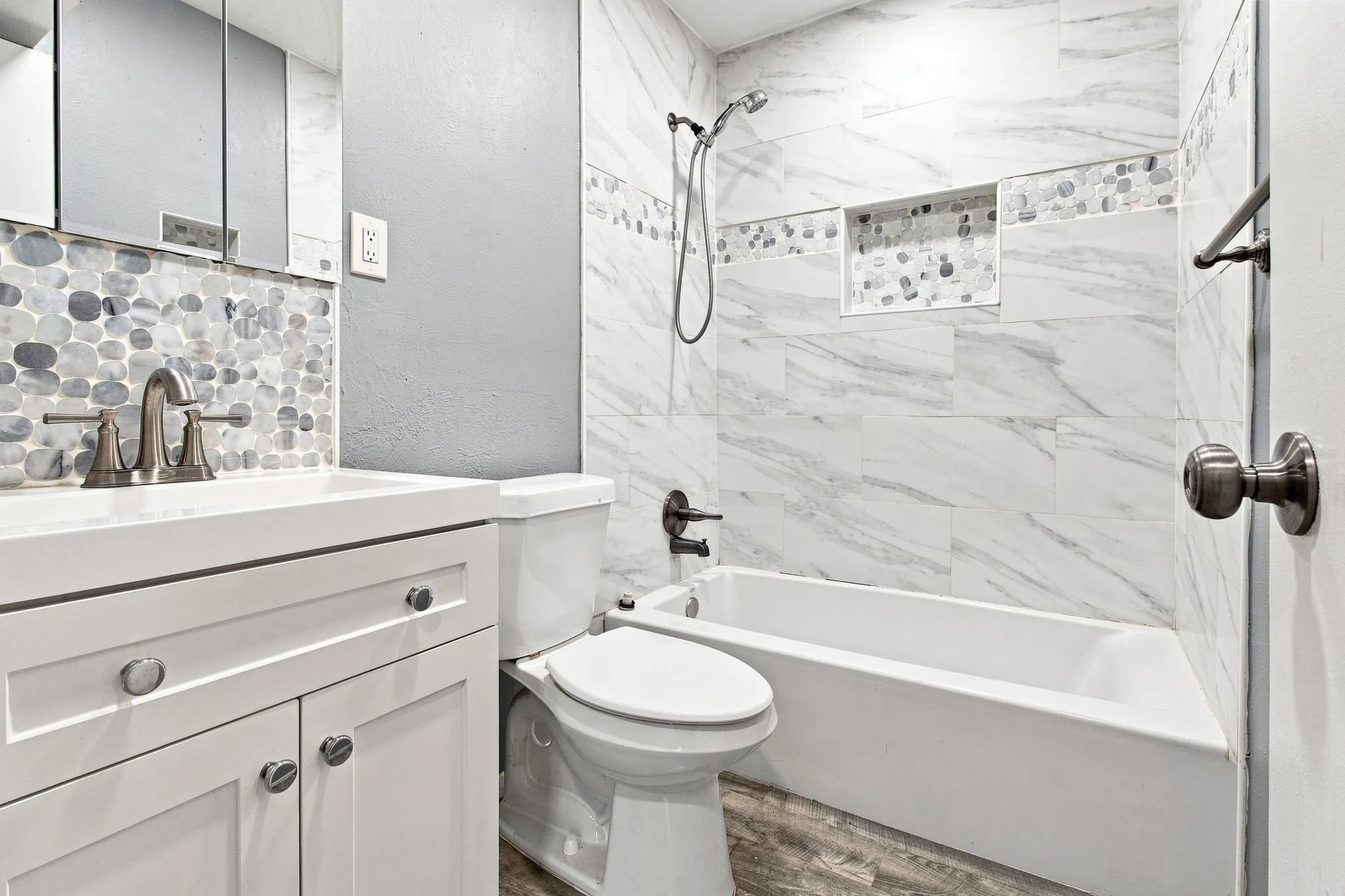 White vanity with brushed nickel hardware, pebble tile backsplash, white tile shower surround with mosaic detail, built-in shower niche, and wood-finish flooring