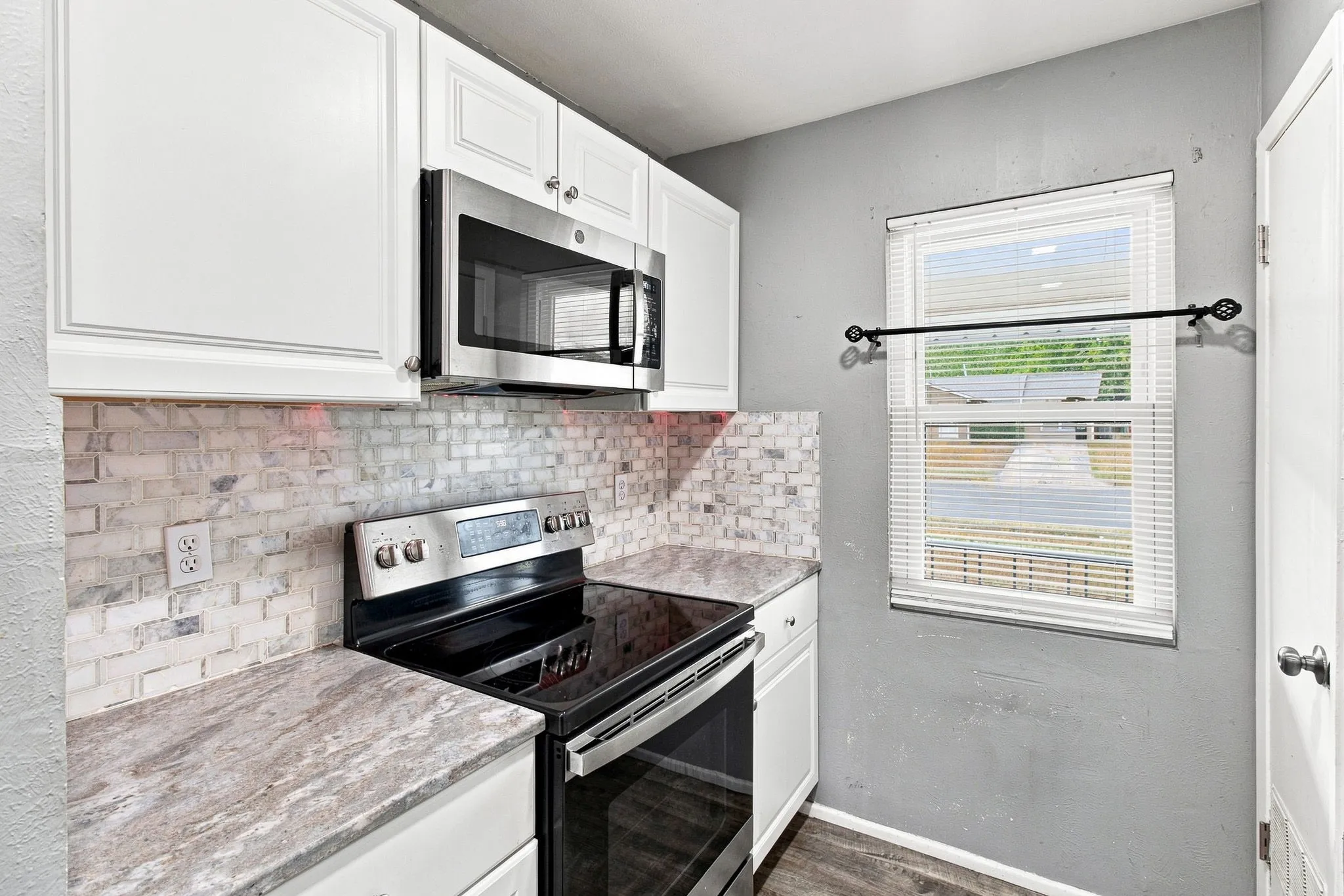 Stainless steel microwave, white upper and lower cabinetry, patterned tile backsplash, solid surface countertop, and wood-finish flooring
