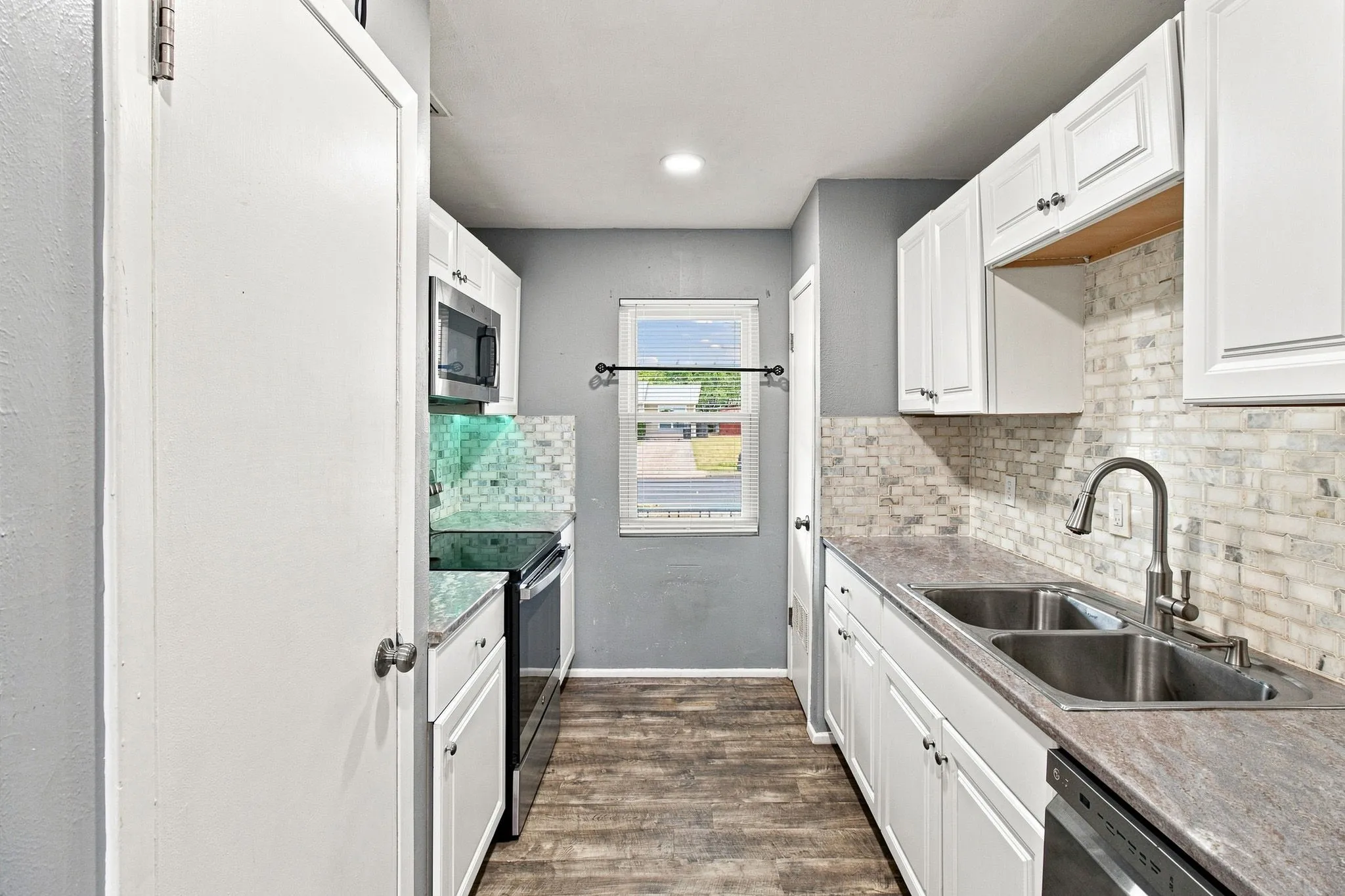 Galley kitchen featuring white cabinetry, wood-finish flooring, a double basin stainless steel sink, and a mosaic tile backsplash