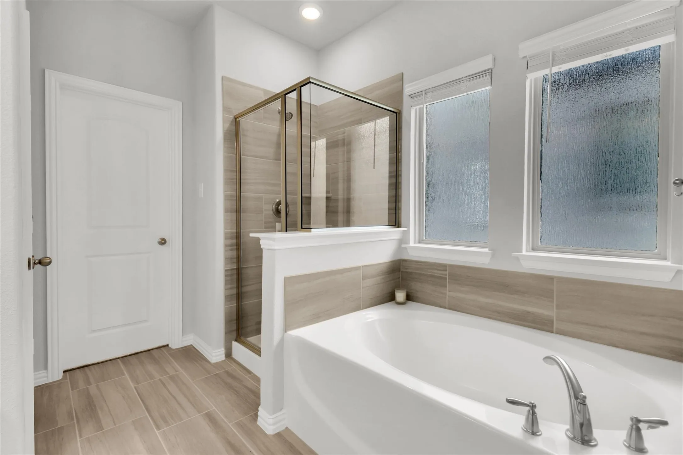 Recessed lighting, large soaking tub with chrome fixtures, tiled shower with glass enclosure, light wood-finish tile flooring, and dual windows with privacy glass