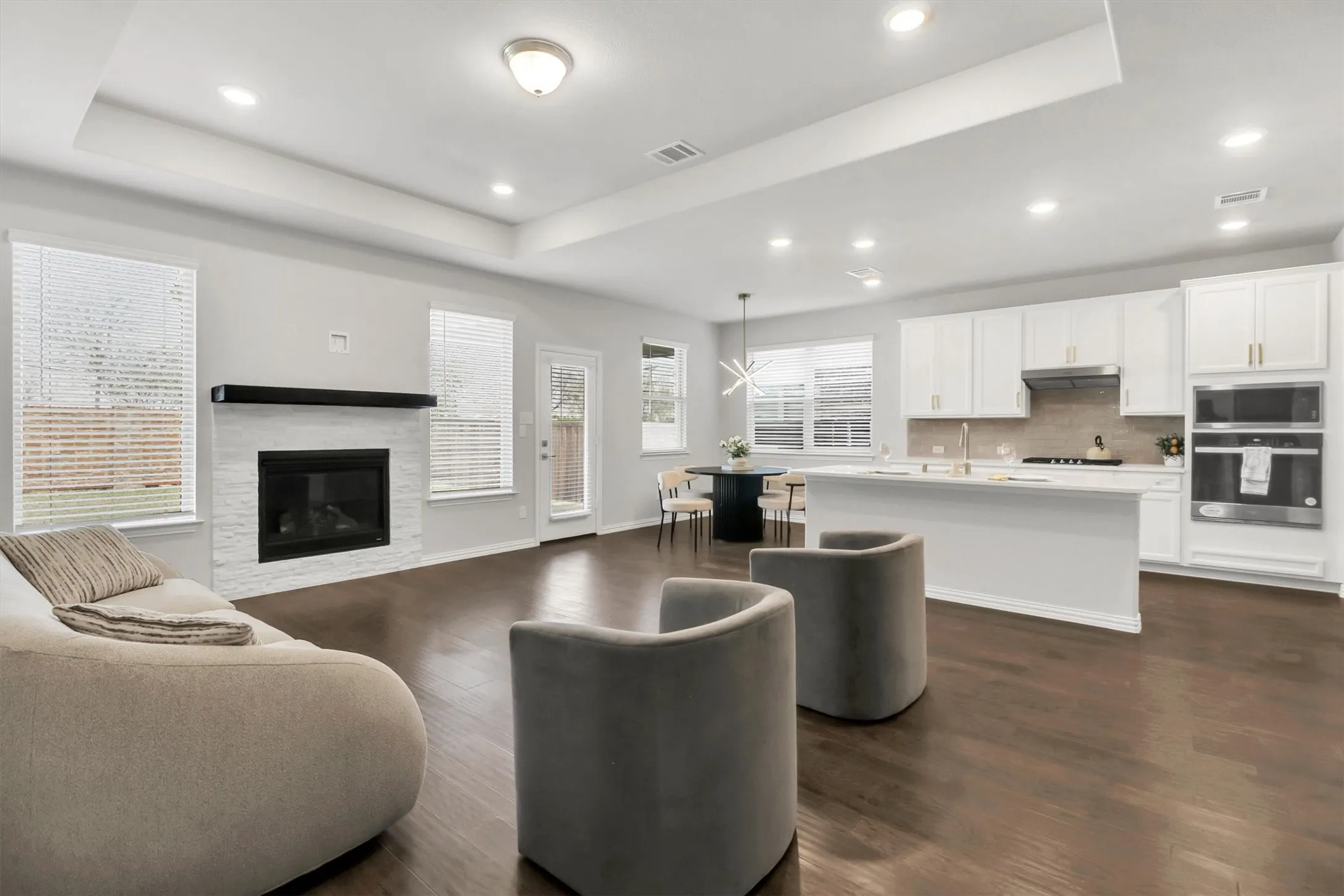 Tray ceiling with recessed lighting, wood-finish flooring, white brick-surround fireplace, white cabinetry with gold-tone hardware, and an island with a sink