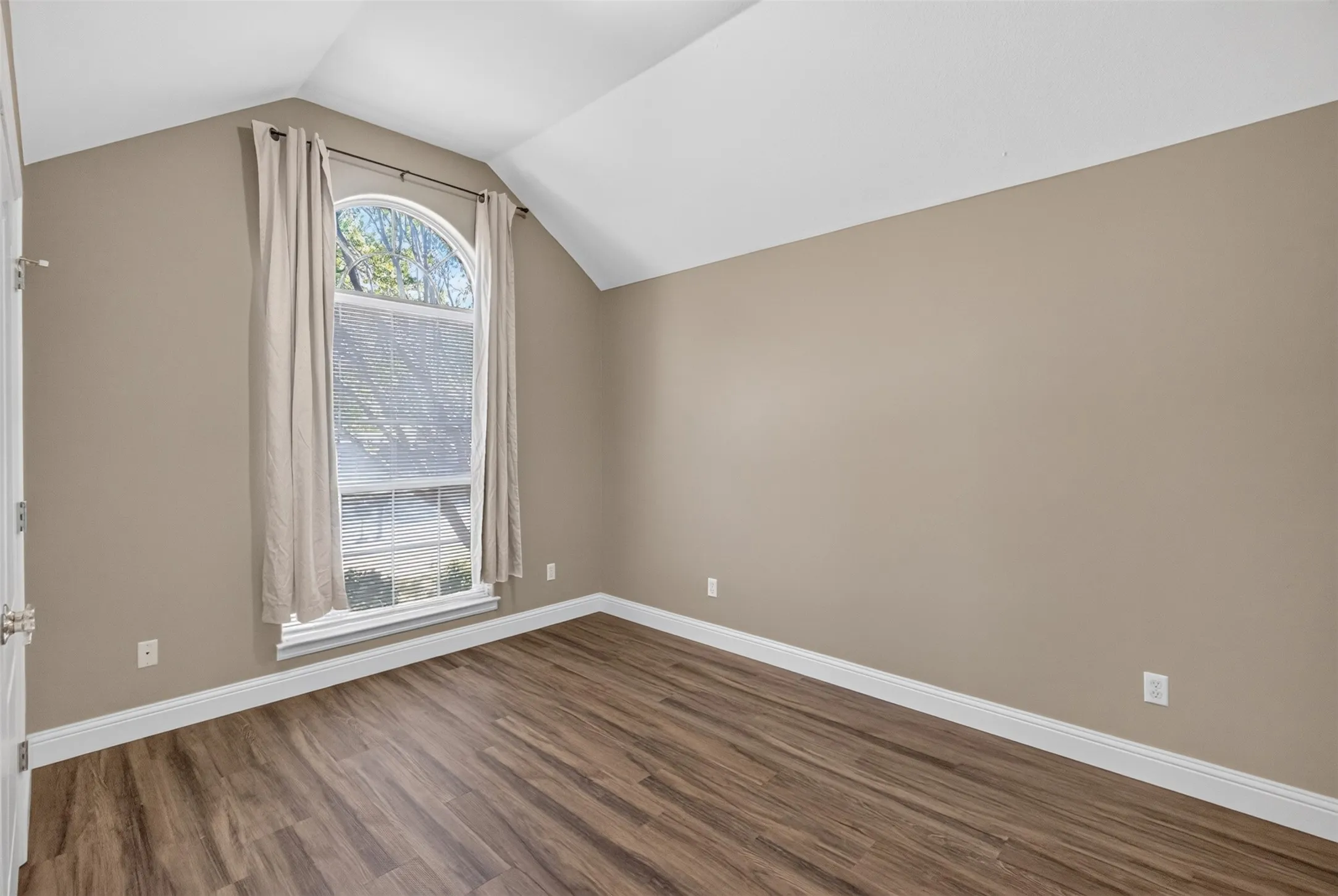 Bright bedroom featuring sloped vaulted ceilings, arched transom window with natural light, wood-finish flooring, and crisp white trim throughout.