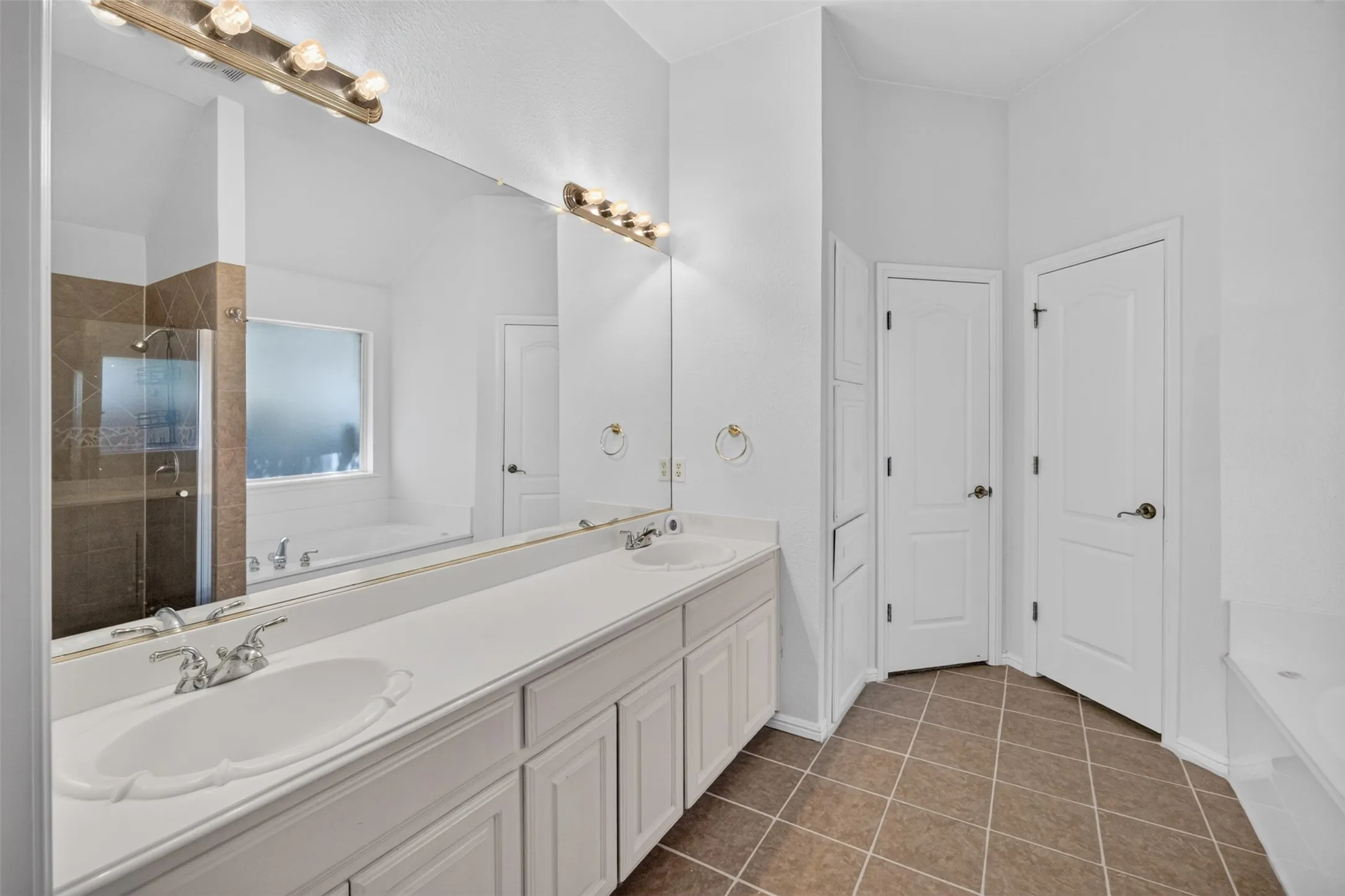 Dual vanity with integrated sinks, expansive wall-mounted mirror, tiled flooring, glass-enclosed shower, and built-in storage