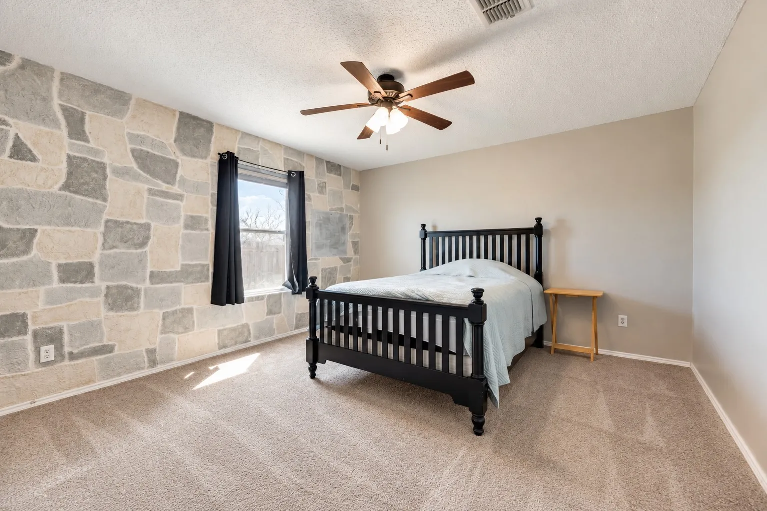 Textured accent wall, ceiling fan with light fixture, window with drapes, and neutral carpet flooring