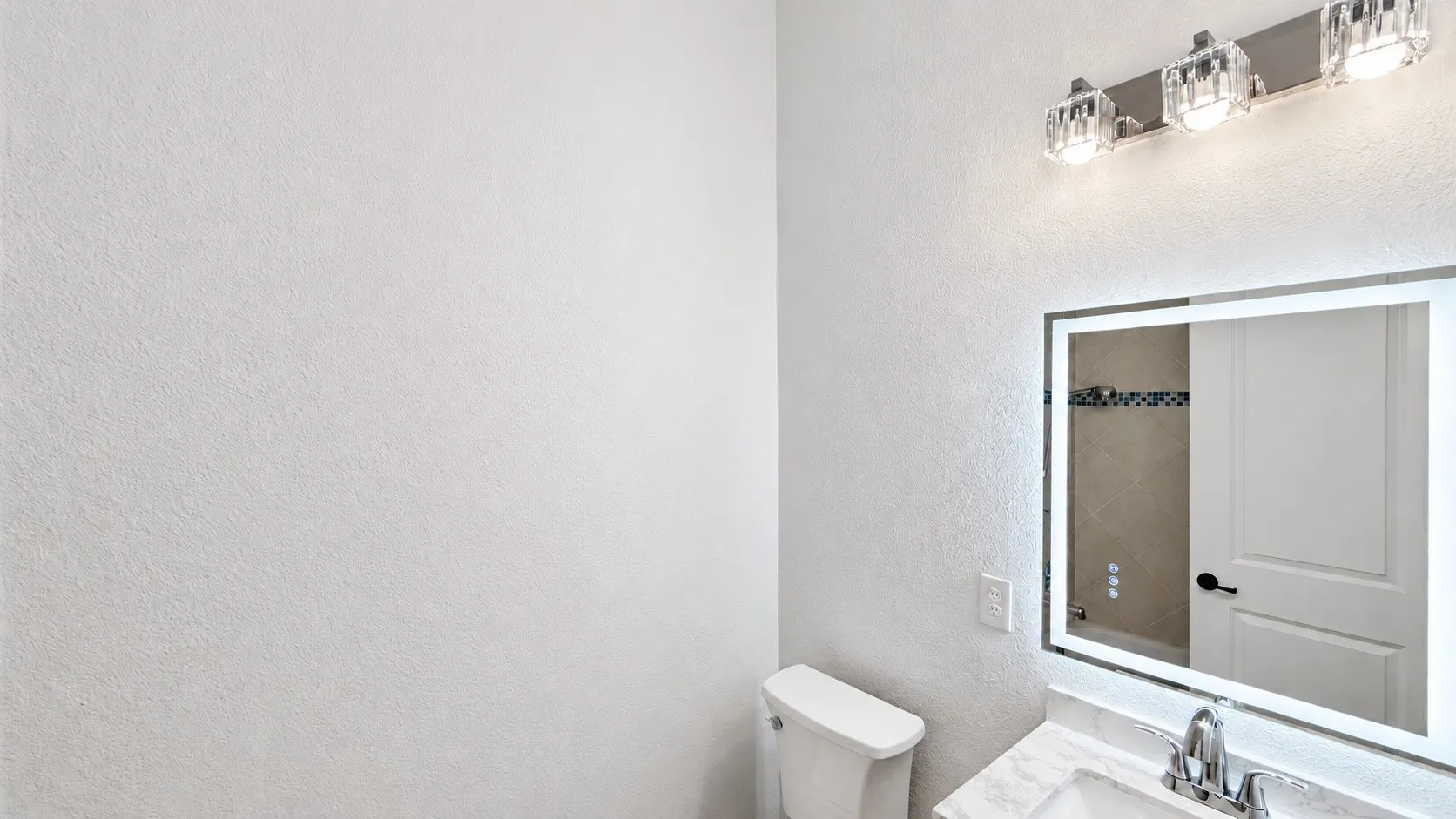 Backlit vanity mirror, modern chrome faucet, white toilet, crystal-accented light fixture, and textured wall finish
