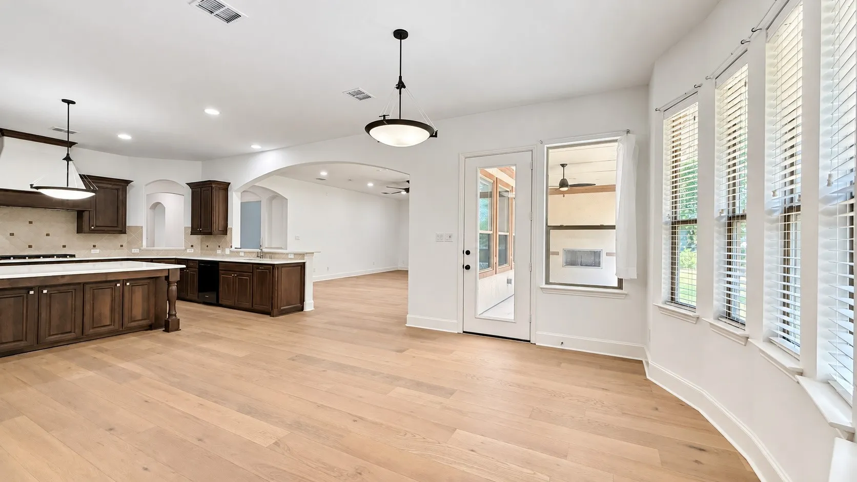 Wood-finish flooring throughout, dark wood cabinetry with light countertops, kitchen island with seating, arched doorways, and a curved window with horizontal blinds