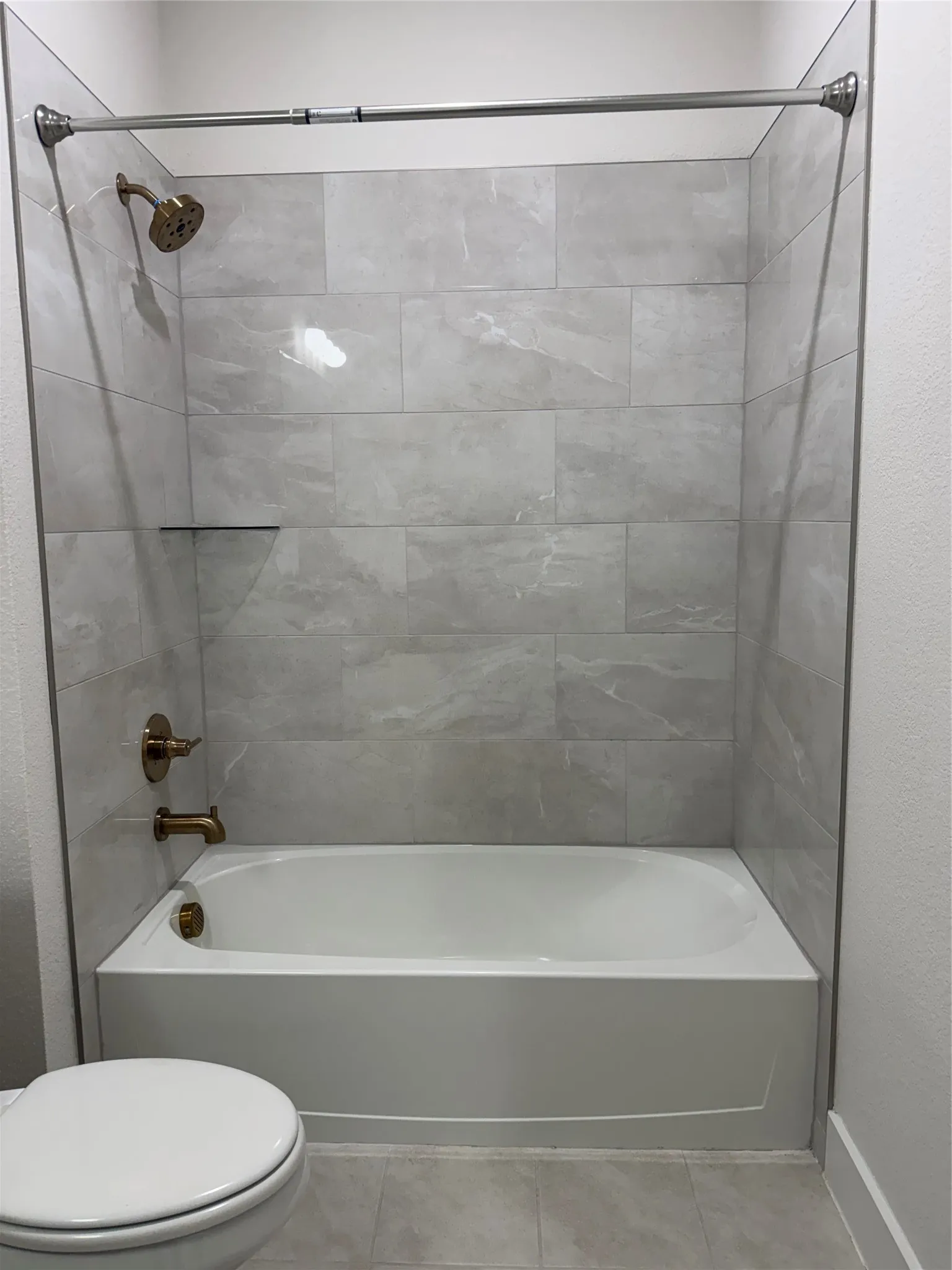 Bathtub with a shower, light grey wall tile, bronze shower fixtures, and a built-in shower shelf