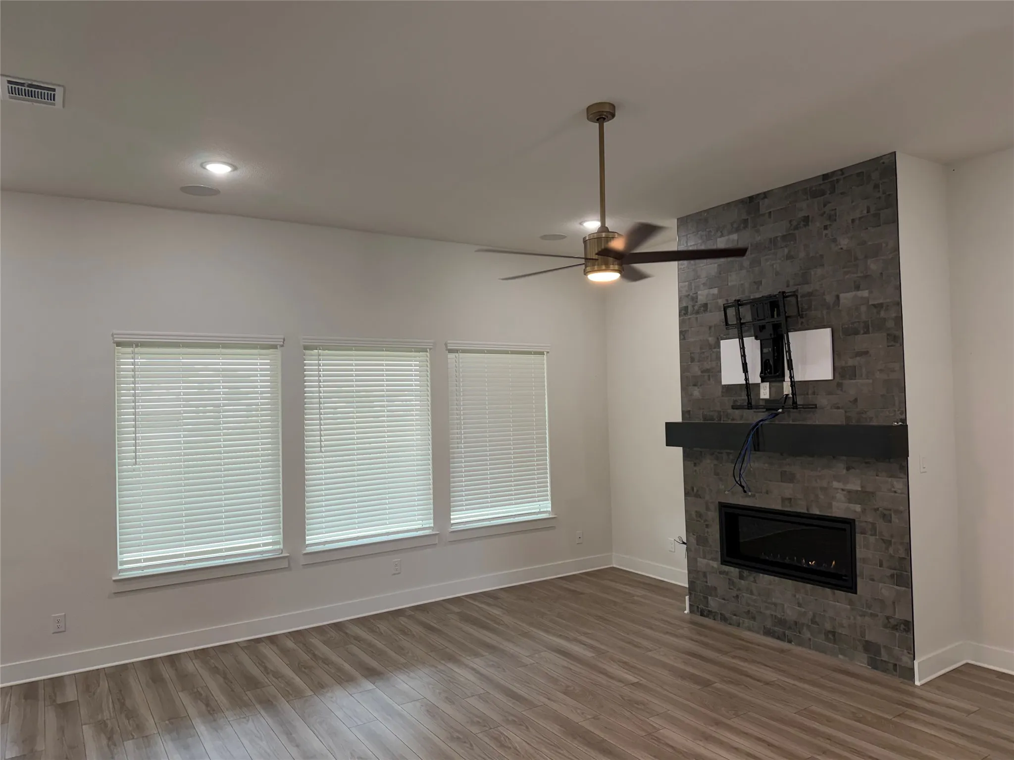 Wood-finish flooring, recessed lighting, ceiling fan, three windows with blinds, and a fireplace with a stone-tile surround and dark mantel