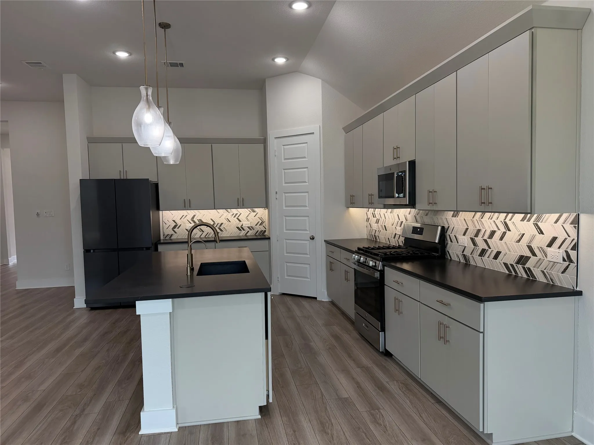 Kitchen featuring sleek gray cabinetry, stainless steel appliances, dark countertops, a chevron tile backsplash, and wood-finish flooring