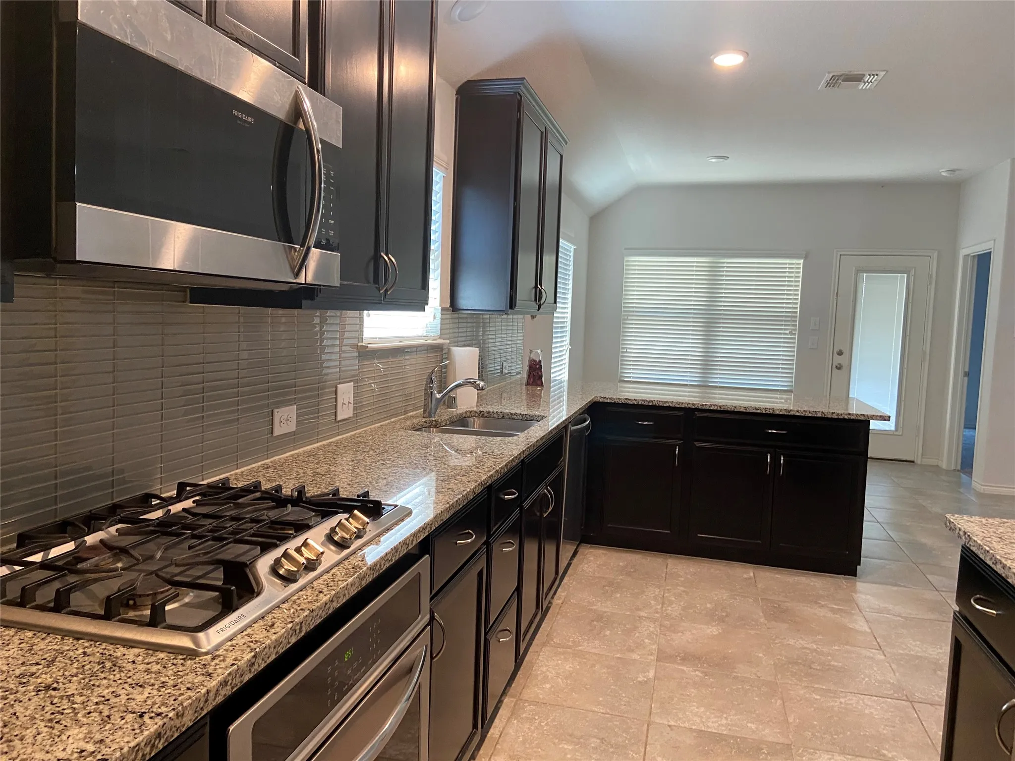 Kitchen with light stone countertops, appliances with stainless steel finishes, tasteful backsplash, dark cabinets, and lofted ceiling