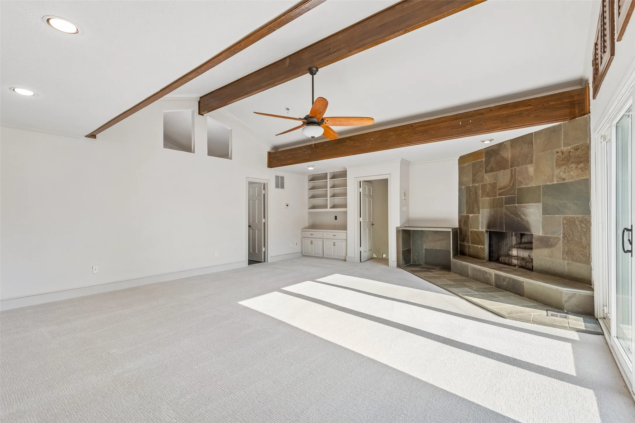 Spacious room featuring exposed ceiling beams, a stone-clad fireplace, integrated shelving, and carpet flooring