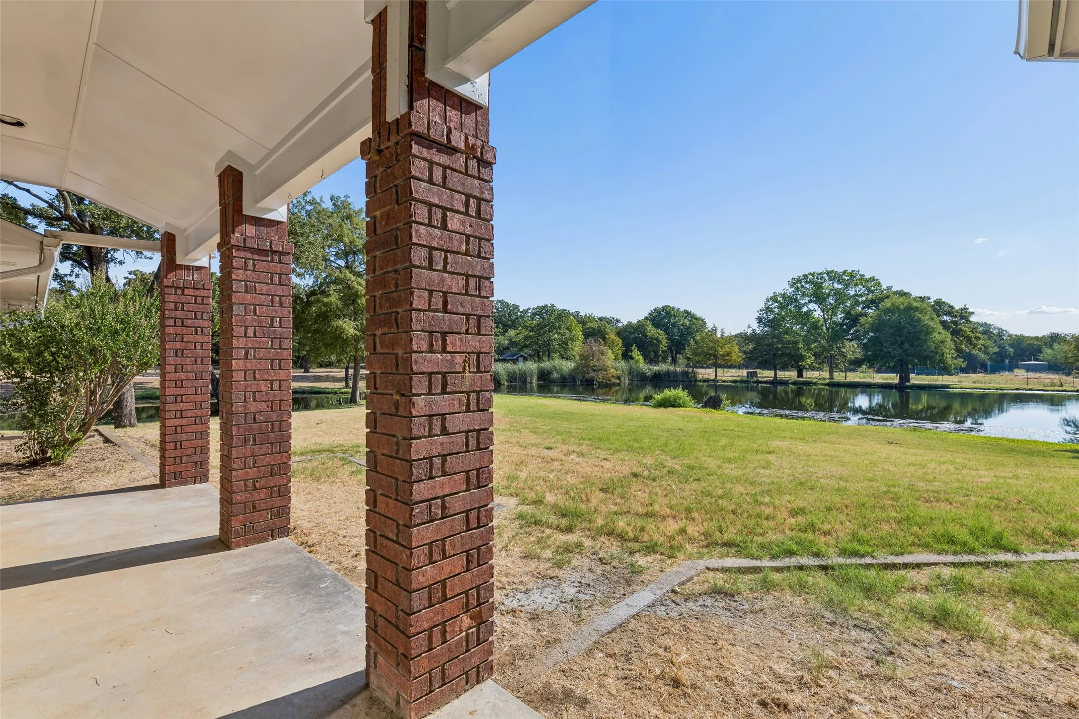 Covered patio with brick columns, concrete flooring, expansive green space, and a prominent water feature