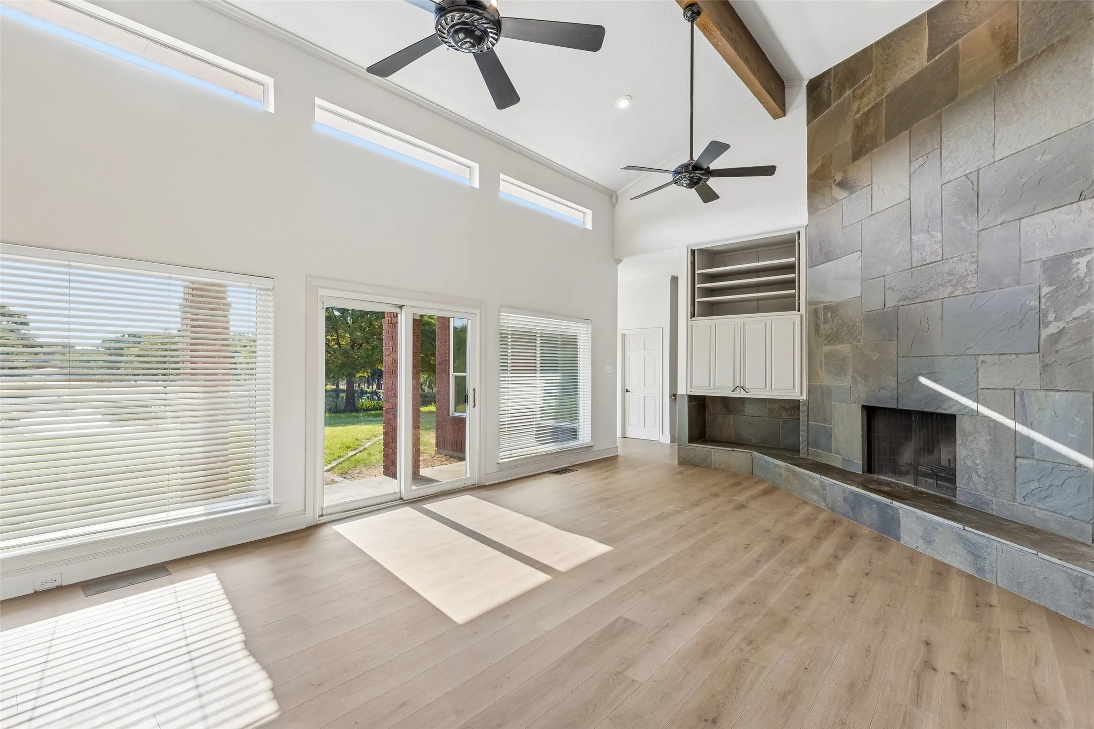 Vaulted ceiling with exposed beam, stone fireplace with hearth, built-in cabinetry, wood-finish flooring, and multiple large windows with sliding glass door