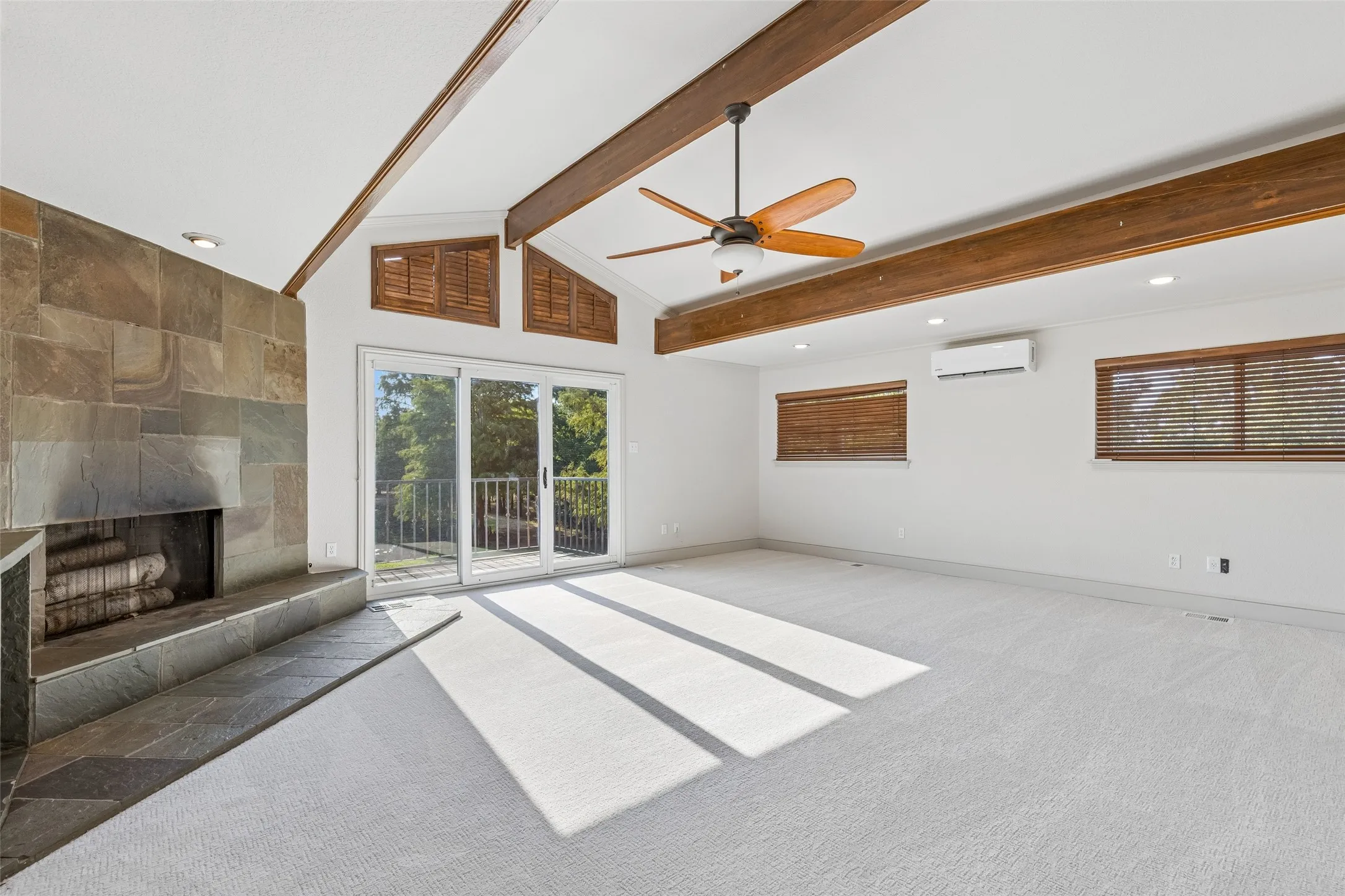 Vaulted ceiling with exposed wood beams, stone-clad fireplace, sliding glass doors to a balcony, ceiling fan, and recessed lighting
