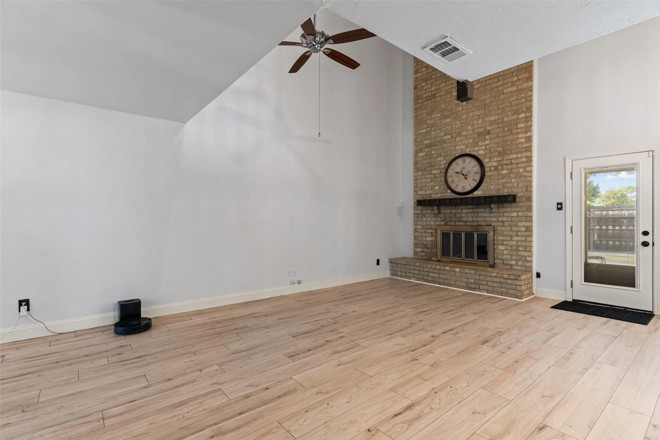 Vaulted ceiling with a ceiling fan, light wood-finish flooring, brick fireplace with a wood mantle, and a glass-paneled door