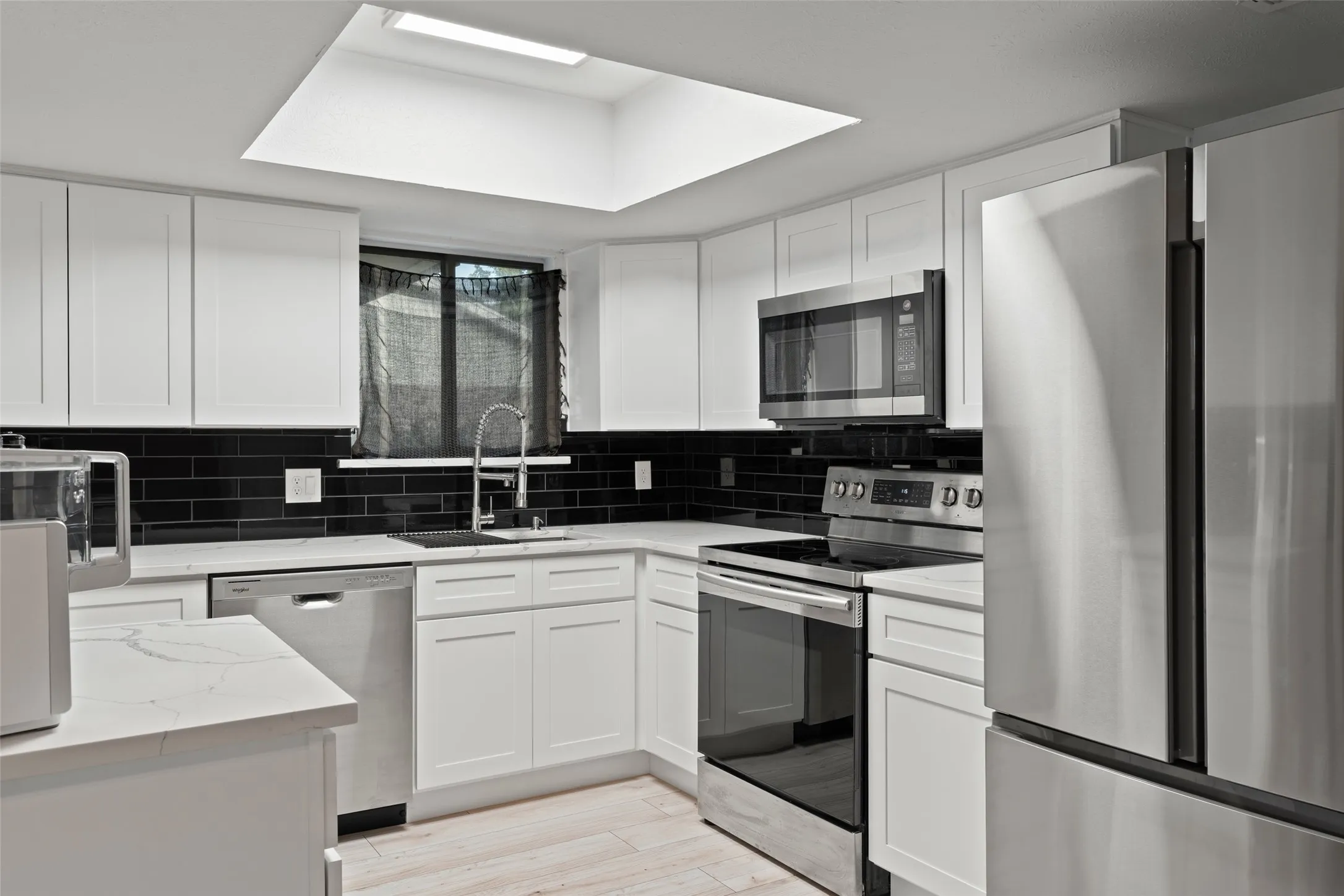 Recessed ceiling lighting with skylight, white shaker-style cabinetry, black subway tile backsplash, stainless steel appliances, and light wood-finish flooring