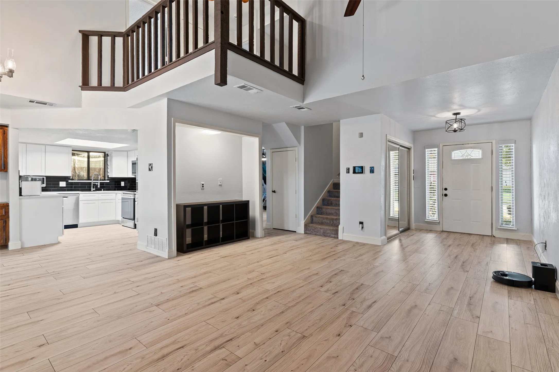 Open-concept living area featuring wood-finish flooring, a two-story ceiling, a contemporary ceiling light fixture, a built-in shelving unit, and a kitchen with white cabinetry and dark backsplash