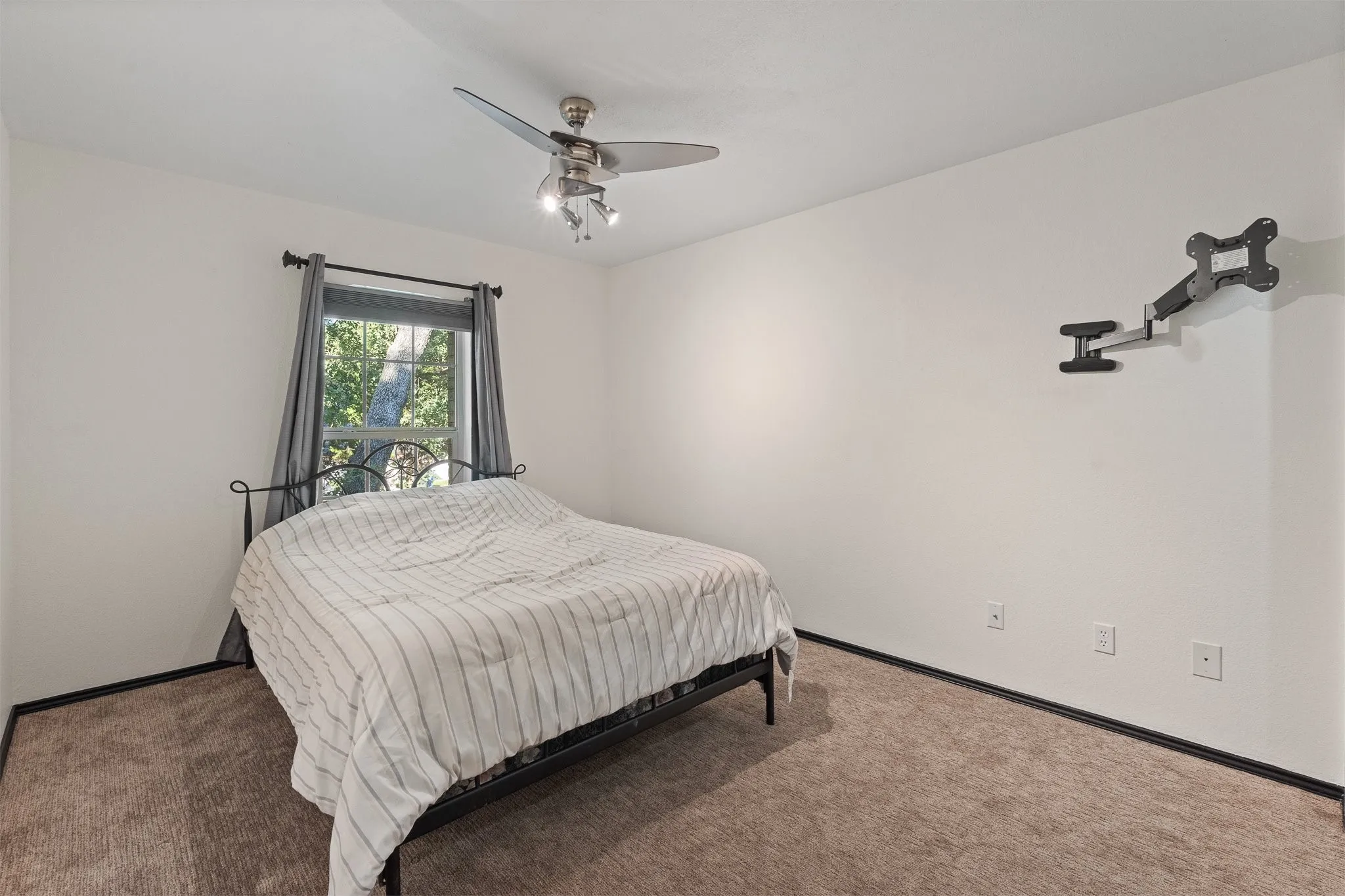 Neutral wall color, window with exterior scenery, ceiling fan with integrated lighting, wall-mounted television bracket, and carpeted flooring