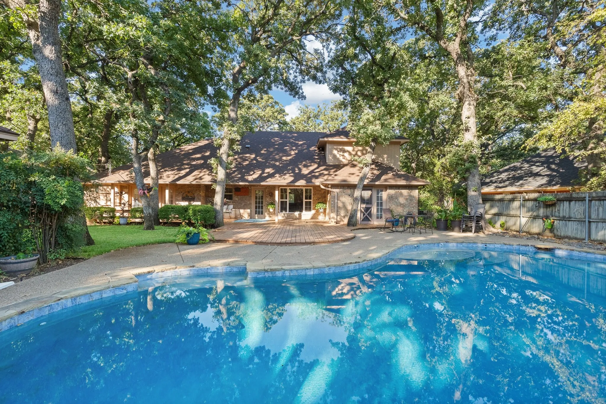 In-ground swimming pool with a surrounding concrete deck, brick paver patio, lush green lawn, and mature trees