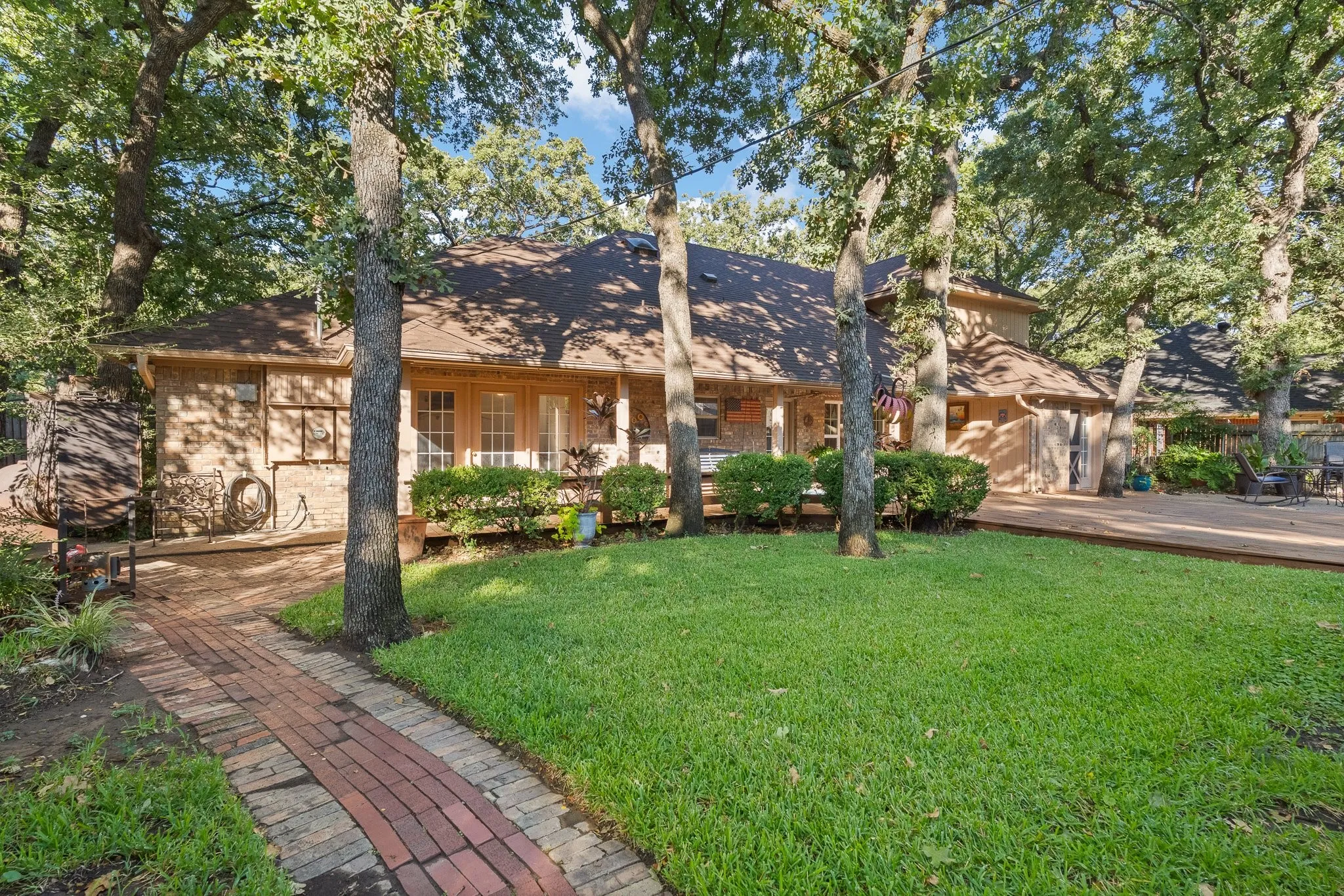 Brick paver walkway leading to a covered porch with column supports, mature trees, green lawn, and an expansive wood deck