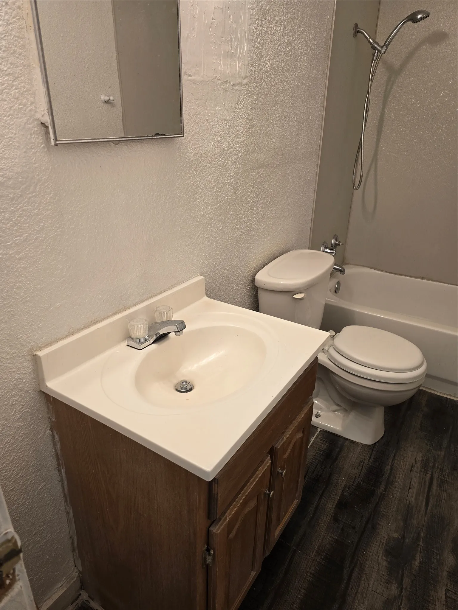 Wood-finish flooring, vanity with integrated sink, chrome faucet, toilet, and bathtub with shower fixture