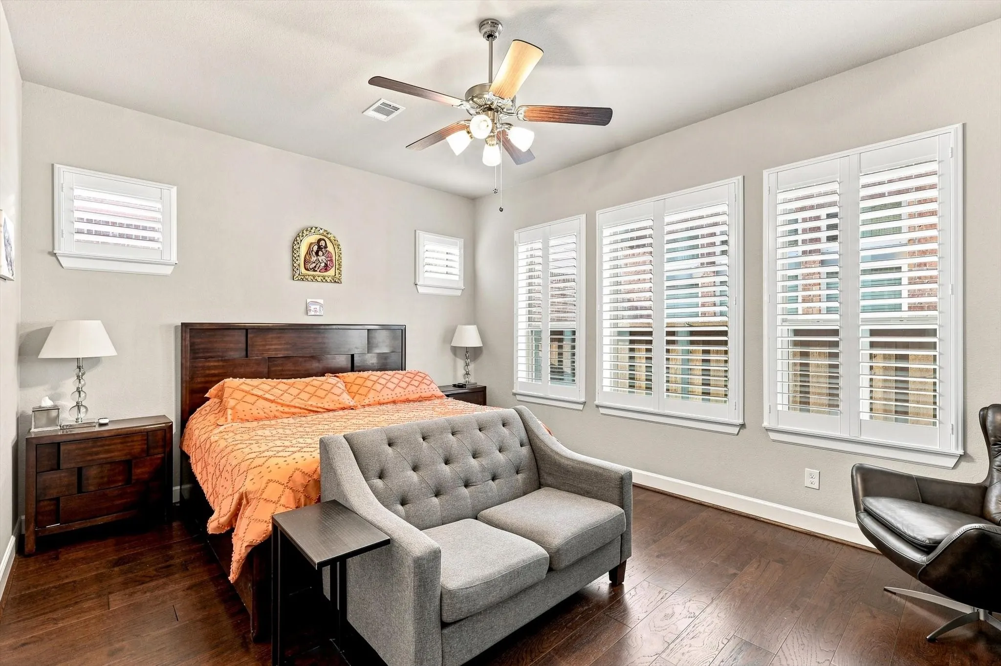 Wood-finish flooring, multiple windows with white shutters, ceiling fan with integrated lighting, and a wood-paneled headboard