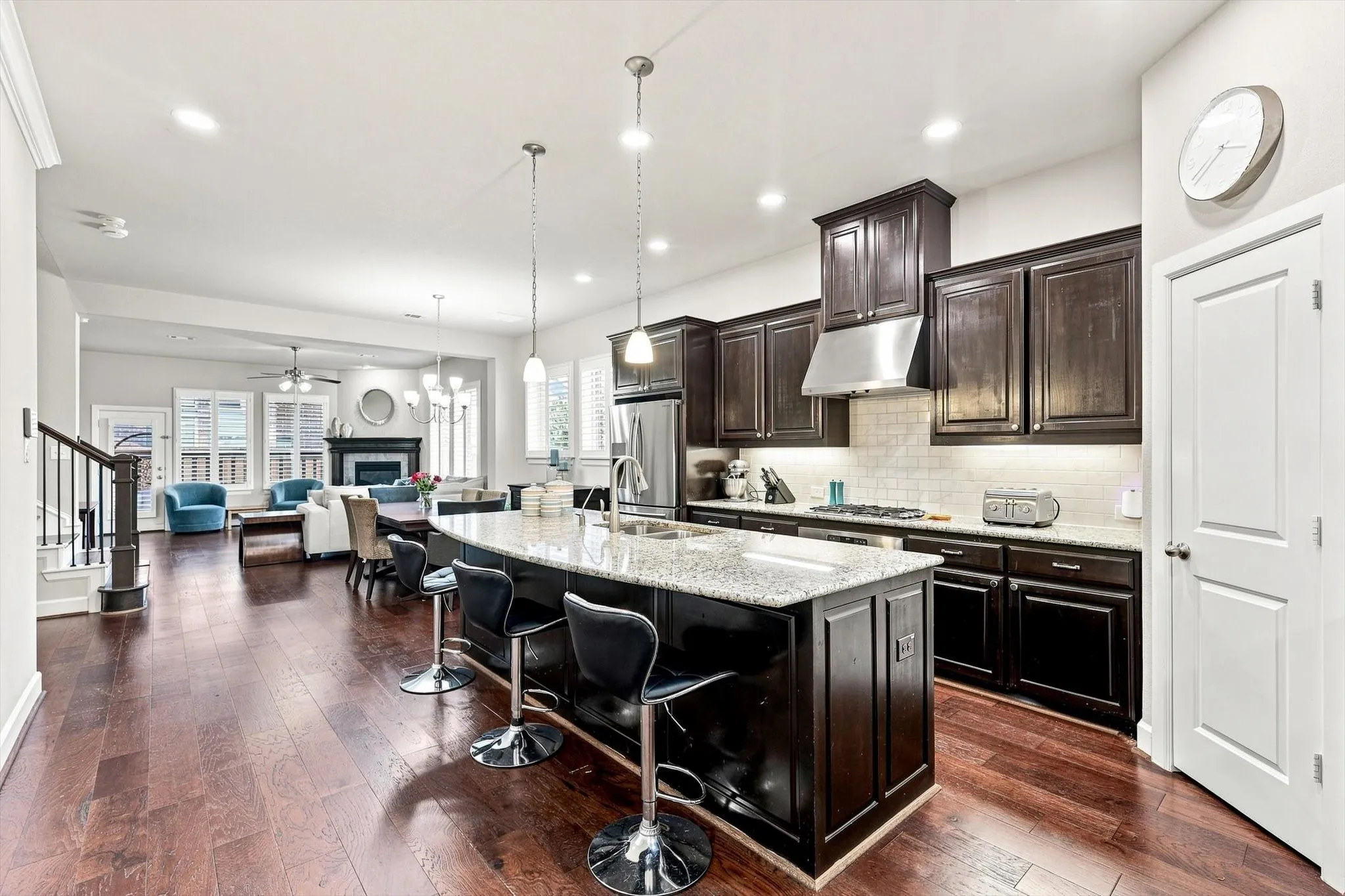Expansive wood-finish flooring throughout, kitchen island with seating, stainless steel appliances, dark wood cabinetry, and a white subway tile backsplash