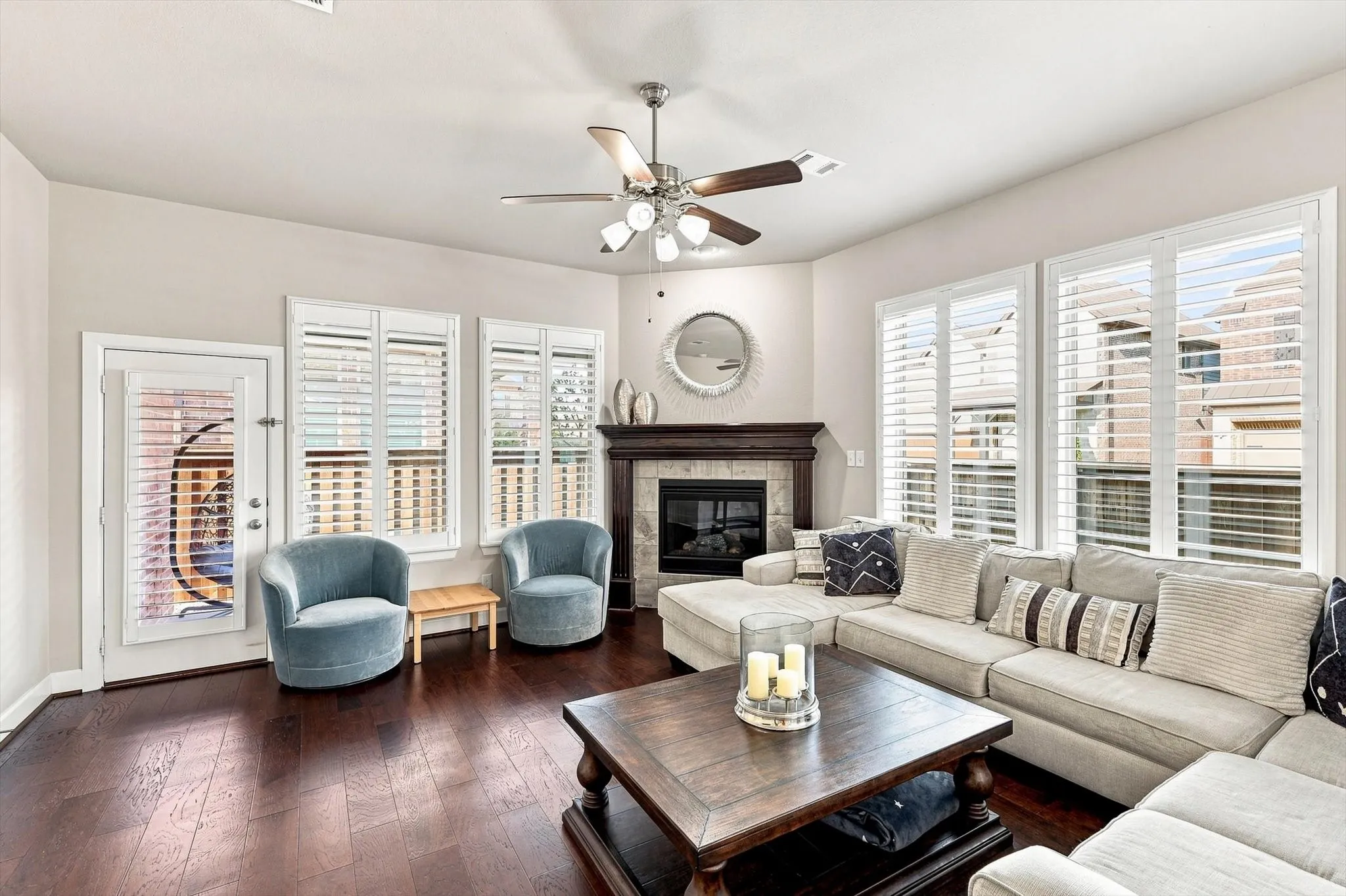 Hardwood flooring throughout, multiple windows with plantation shutters, ceiling fan with integrated lighting, and a corner fireplace with a dark wood mantel and tile surround