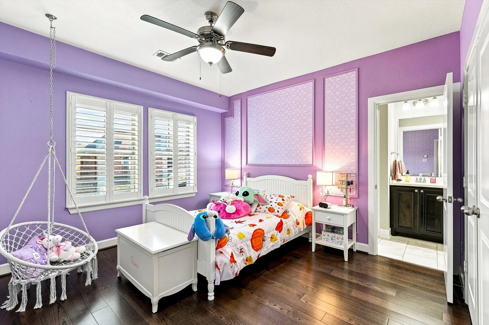 Wood-finish flooring, two windows with white louvered shutters, ceiling fan with integrated lighting, decorative wall panels, and an attached full bathroom with a dark wood-finish vanity
