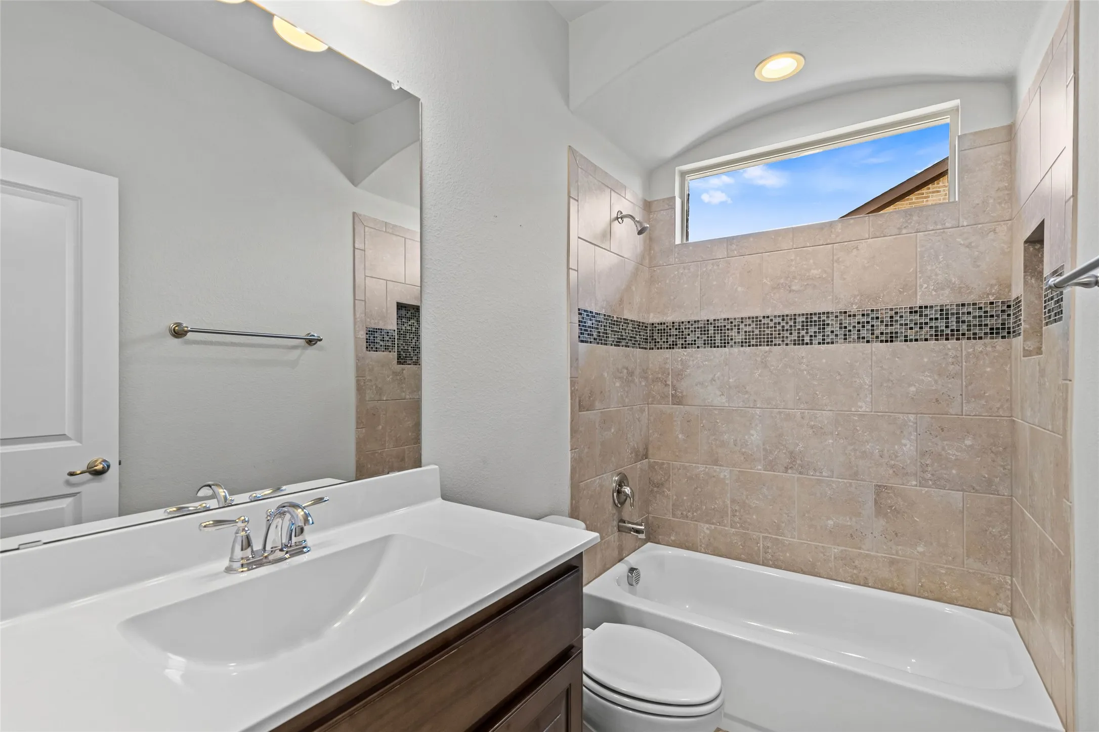 Bathroom featuring a built-in bathtub, a tiled shower surround with an accent mosaic band, an arched window, a vanity with a white countertop and undermount sink, and a large wall-mounted mirror