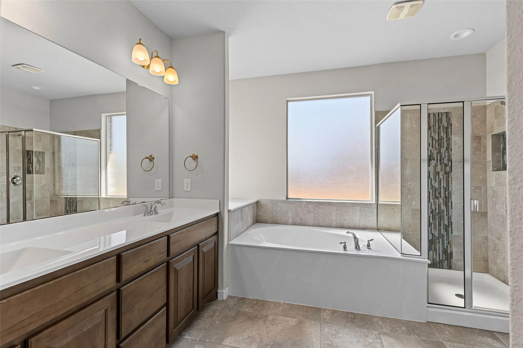 Dual vanity with integrated sinks, wood-finish cabinetry, built-in soaking tub, tile shower with glass enclosure, and tiled flooring