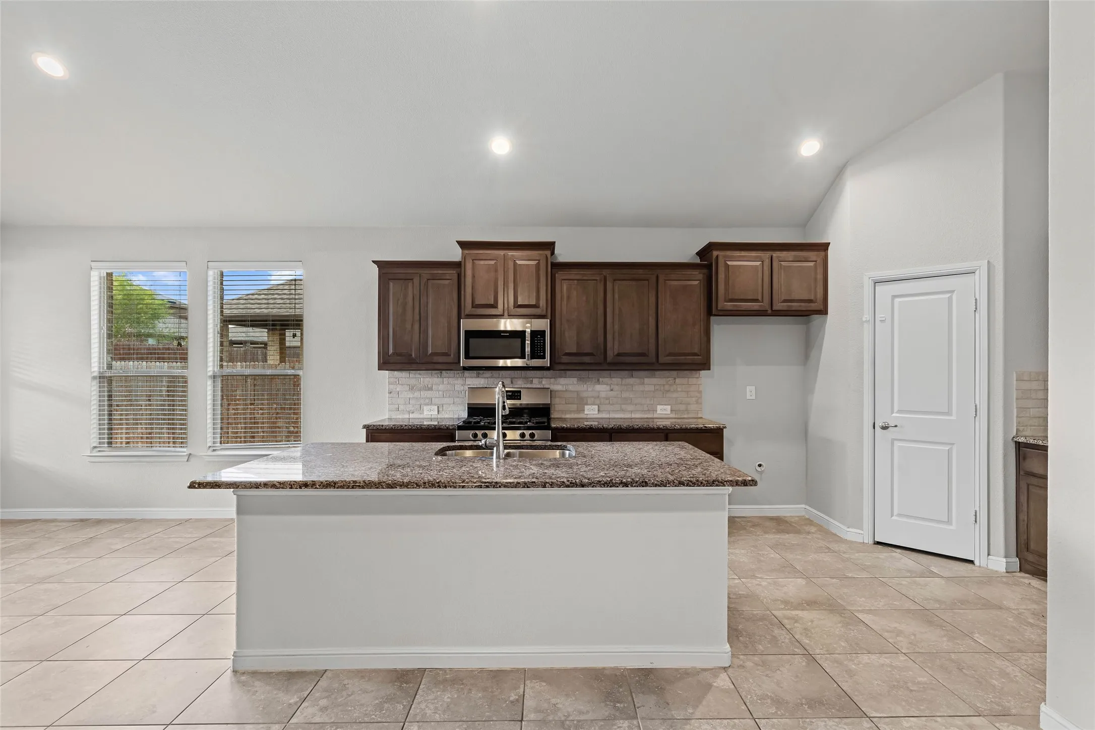 Kitchen featuring an island with a granite-style countertop, dark wood-finish cabinetry, stainless steel appliances, and a tile backsplash